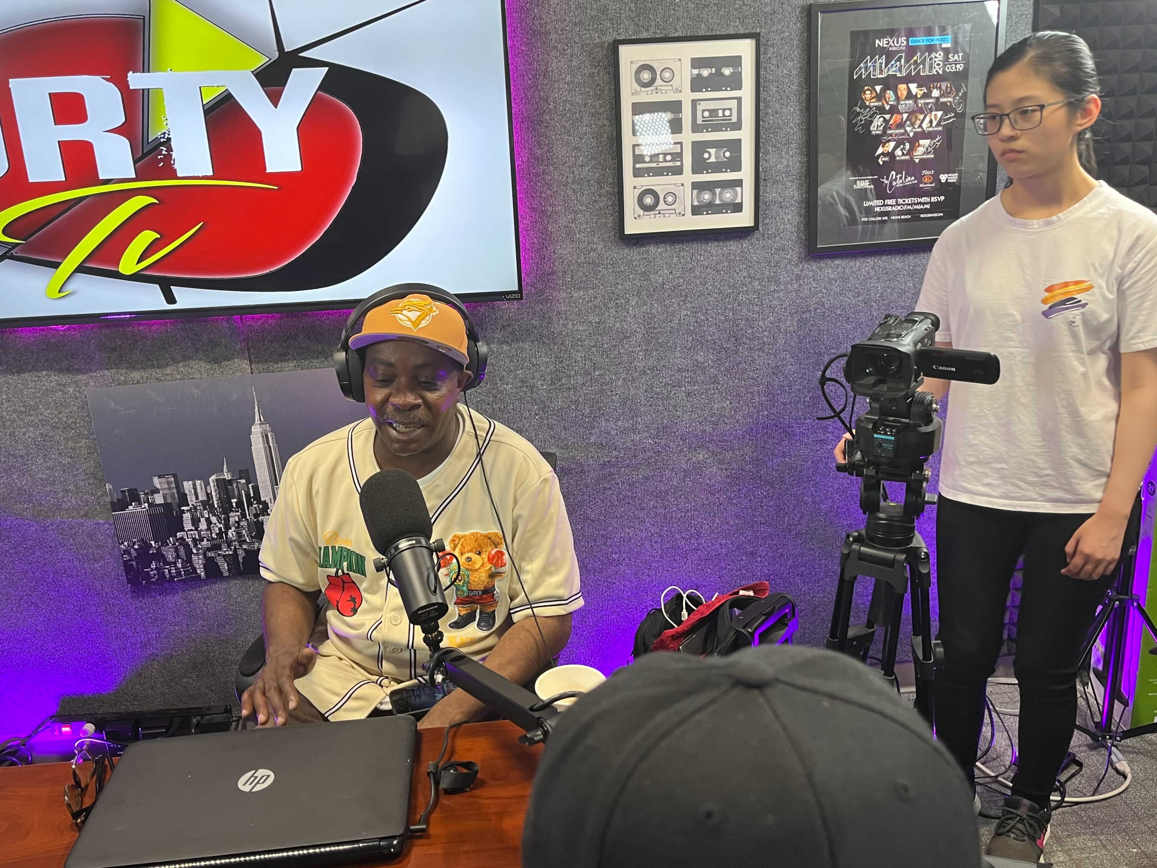 A man in a baseball jersey sits at a desk with a microphone and laptop, while a woman stands nearby operating a camera in a studio setting.