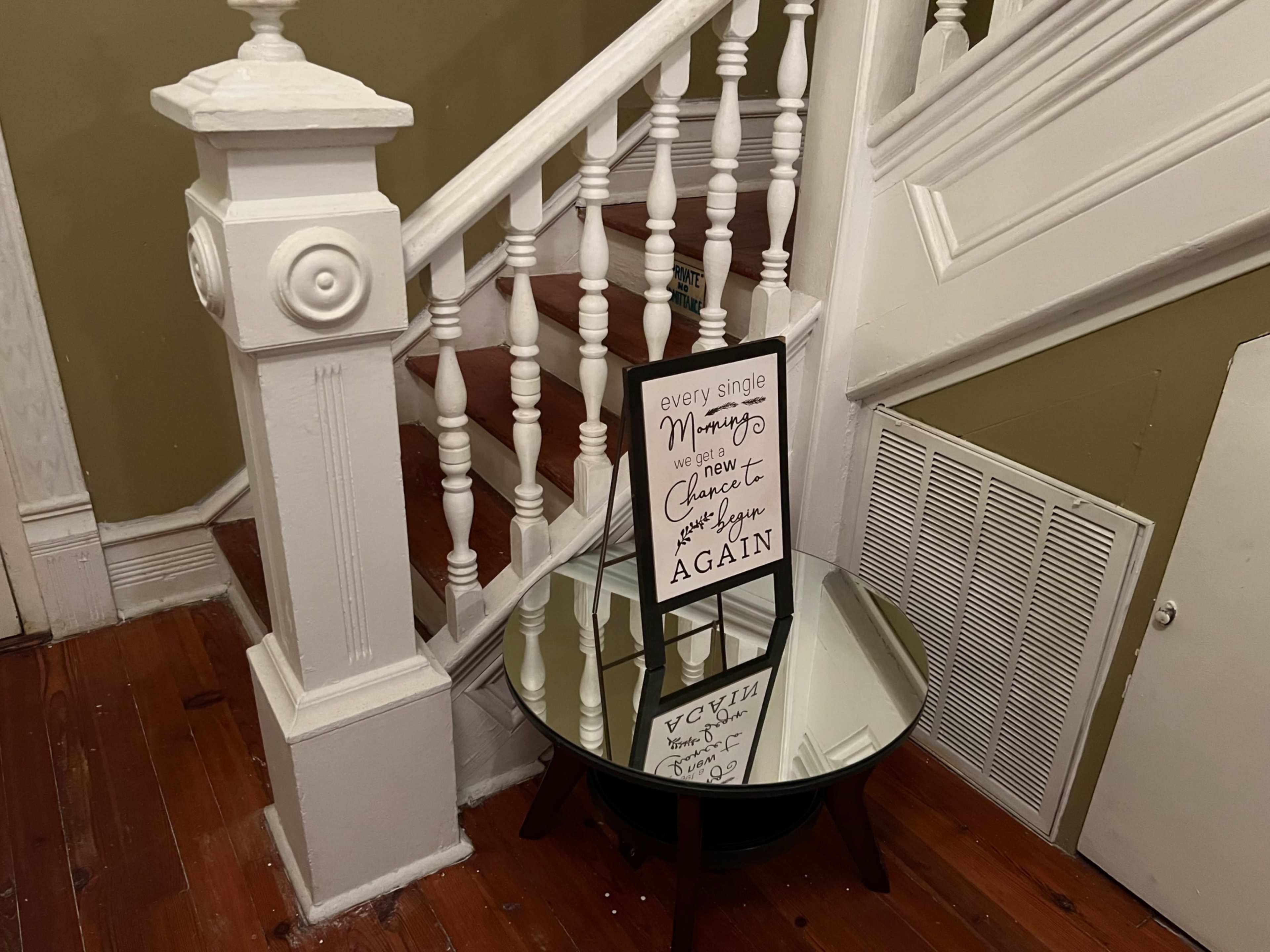 A sign on an easel sits in front of a round mirror table near a staircase with white railings and wooden steps.