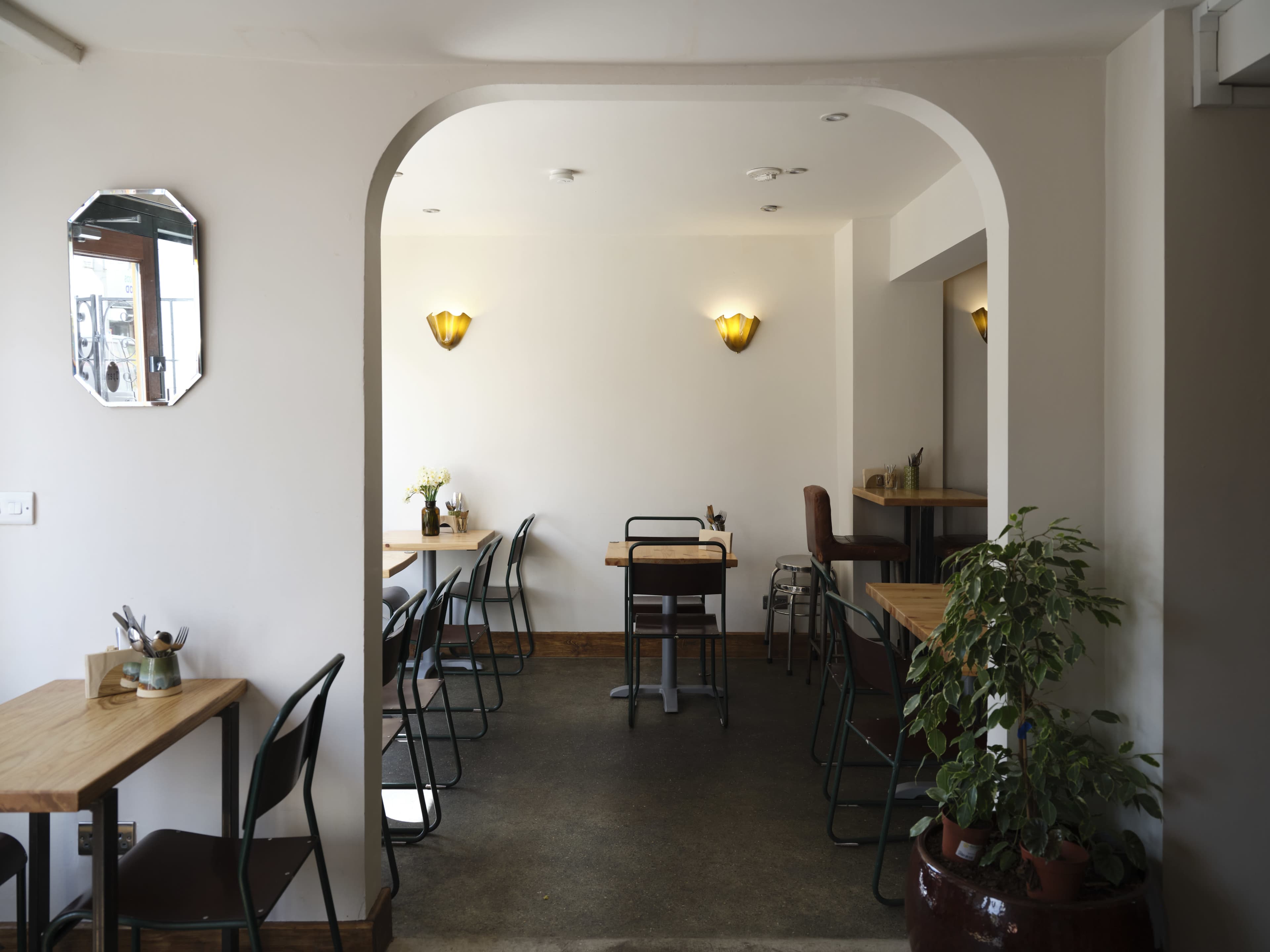 The image shows a minimalist café interior with wooden tables, chairs, and a plant, featuring an archway leading to a well-lit dining area.