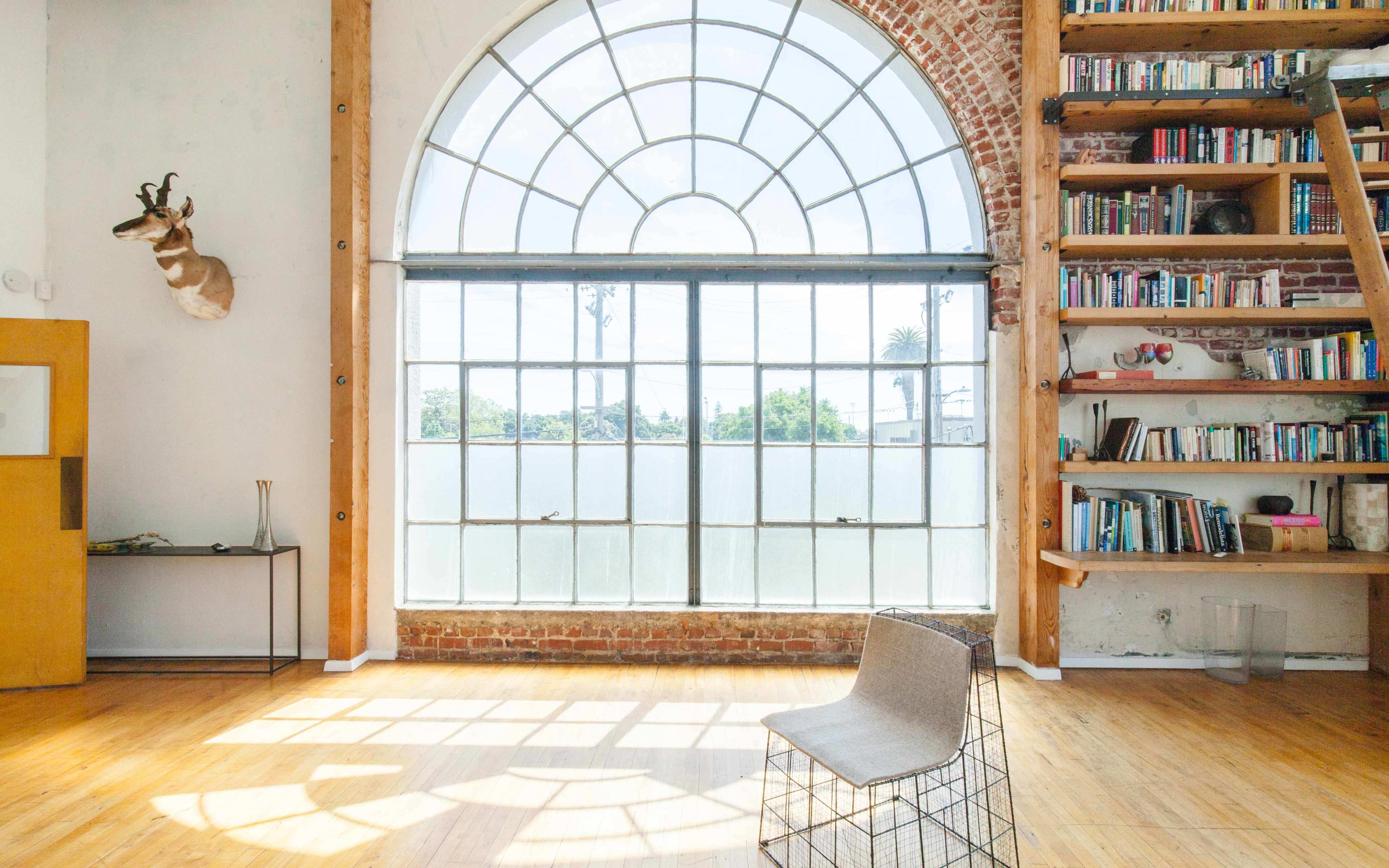 A spacious room features a large arched window, wooden flooring, and a bookshelf against a brick wall, with a single chair positioned near the window.