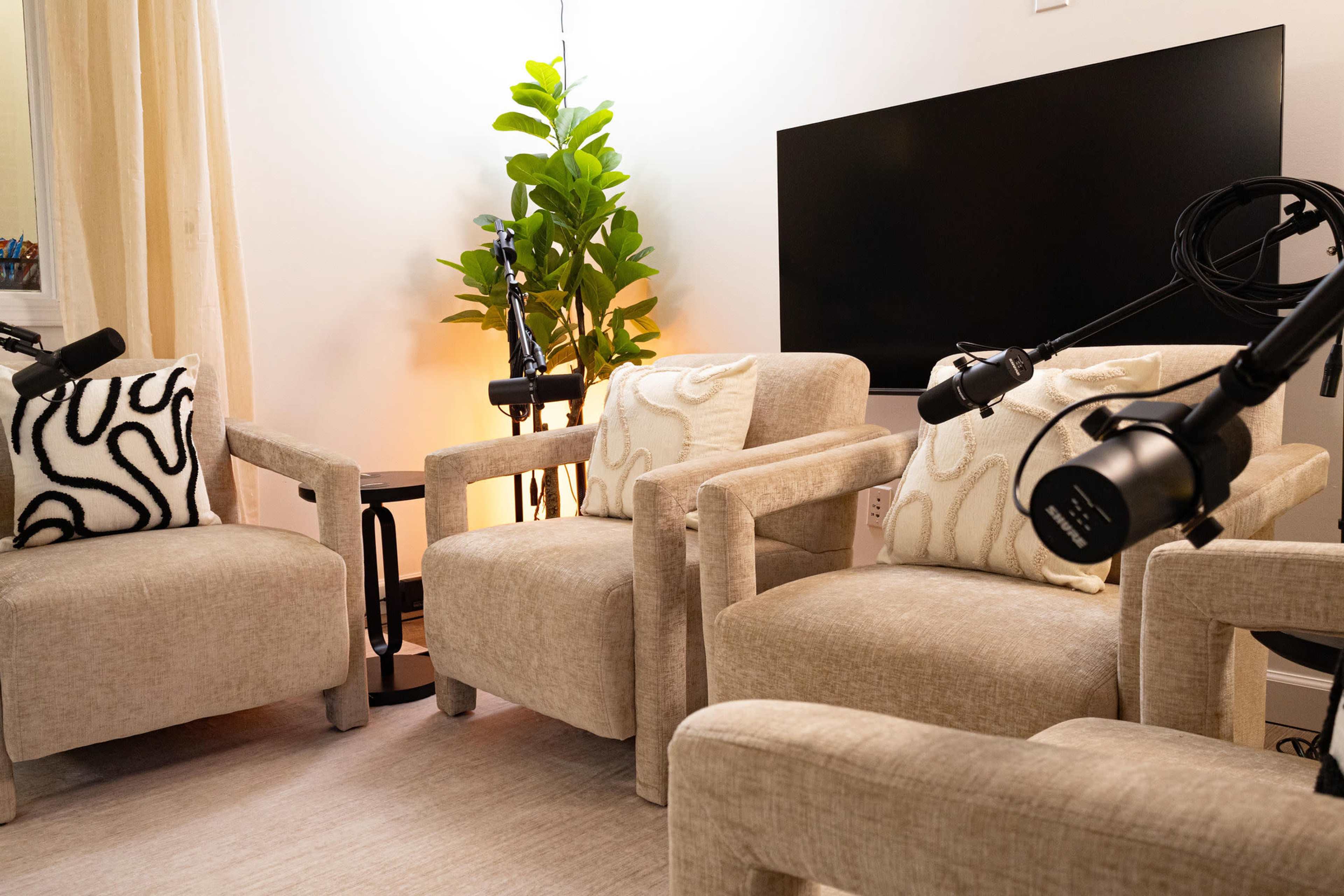 The image shows a cozy conversation area with three beige chairs, a small black table, and two microphones mounted on stands, all set against a simple backdrop of a TV and potted plant.