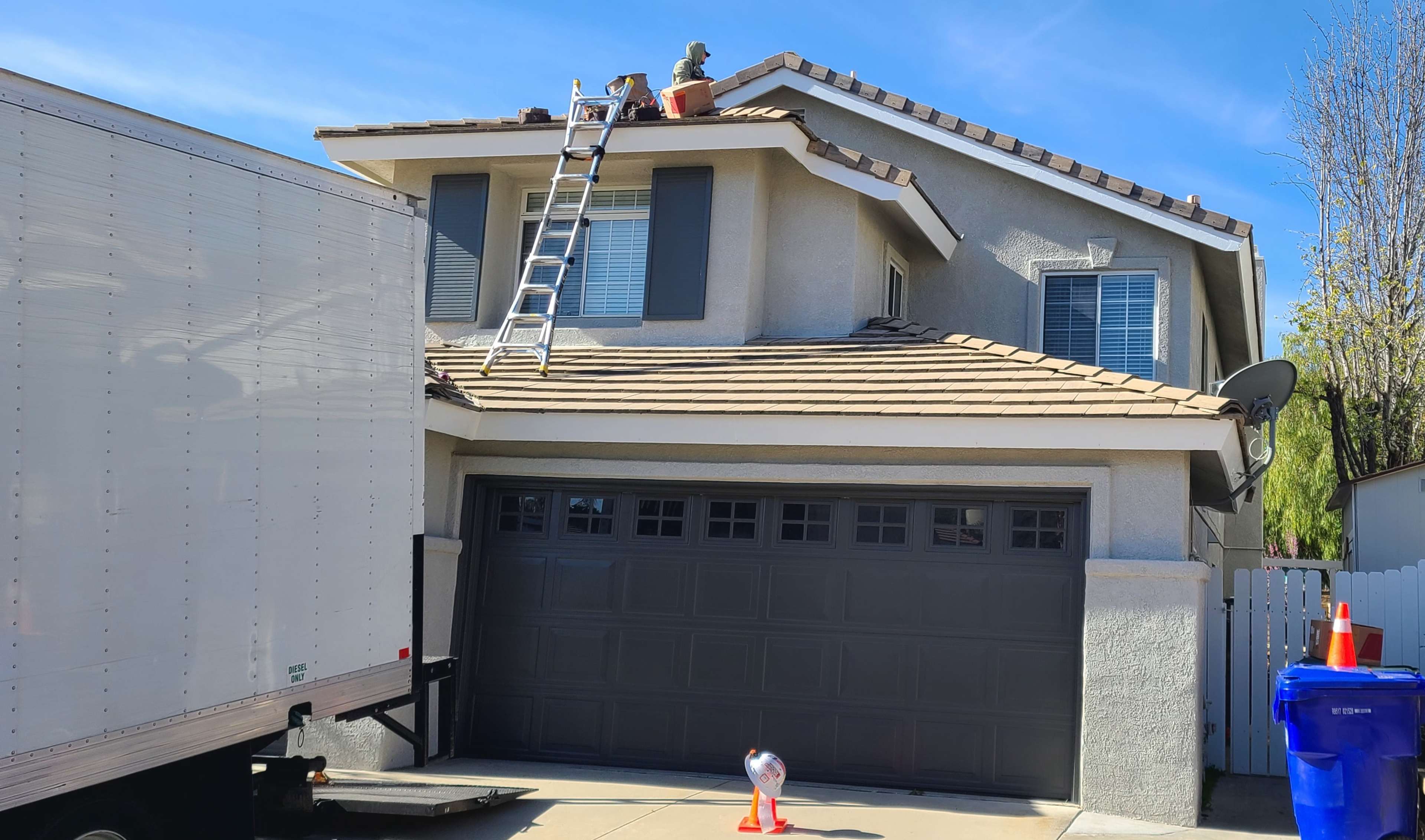 A moving truck is parked in front of a two-story house with a ladder leaning against the roof and a potted plant on top.