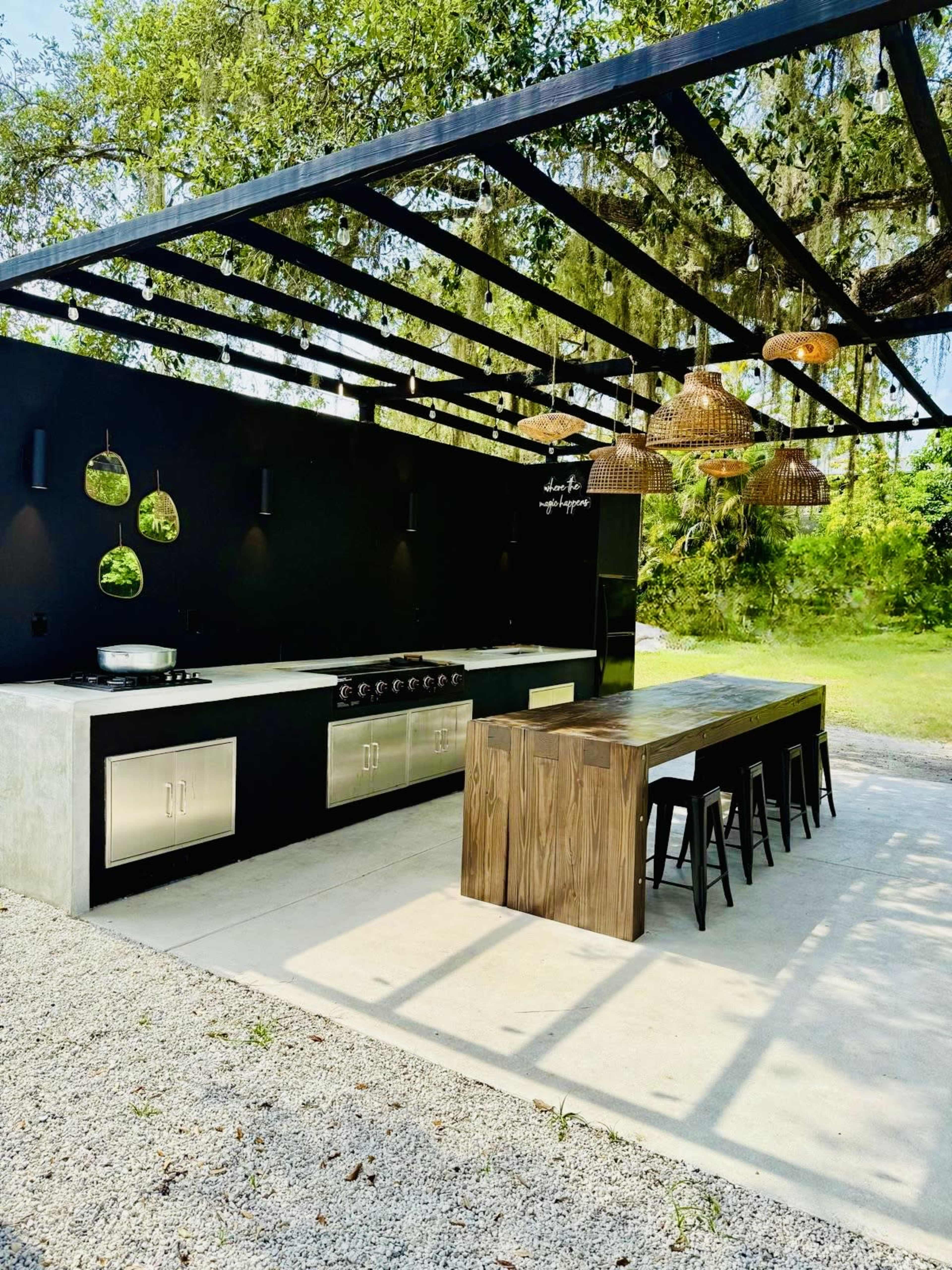The image shows an outdoor kitchen with a modern design featuring a concrete countertop, stainless steel appliances, a wooden dining table, and overhead lighting, all surrounded by greenery.