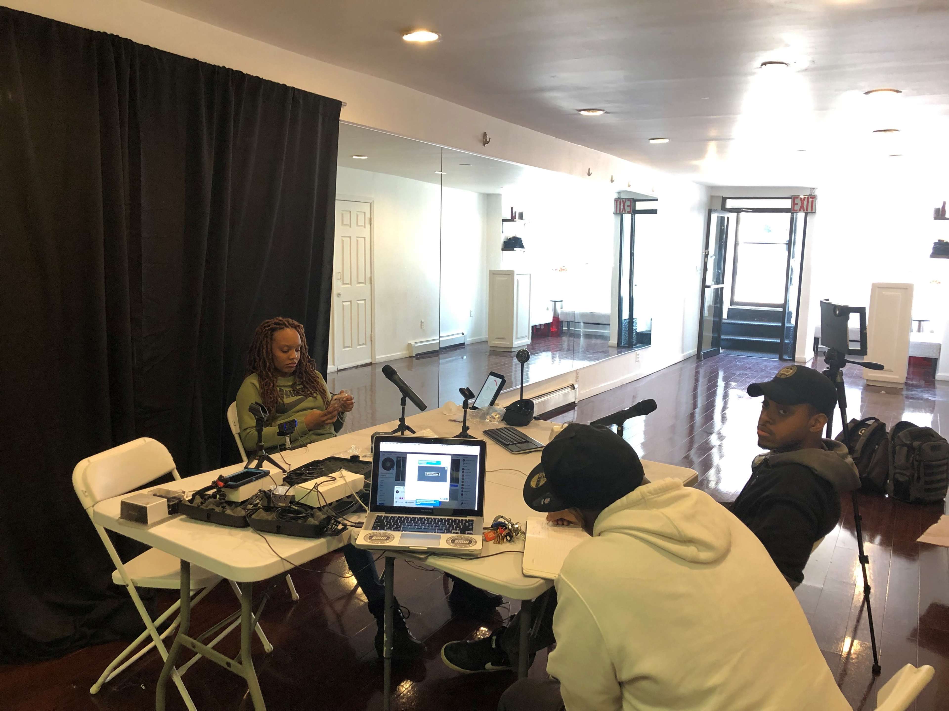 A group of three people is seated around a table with microphones and a laptop in a spacious, well-lit room with mirrored walls.