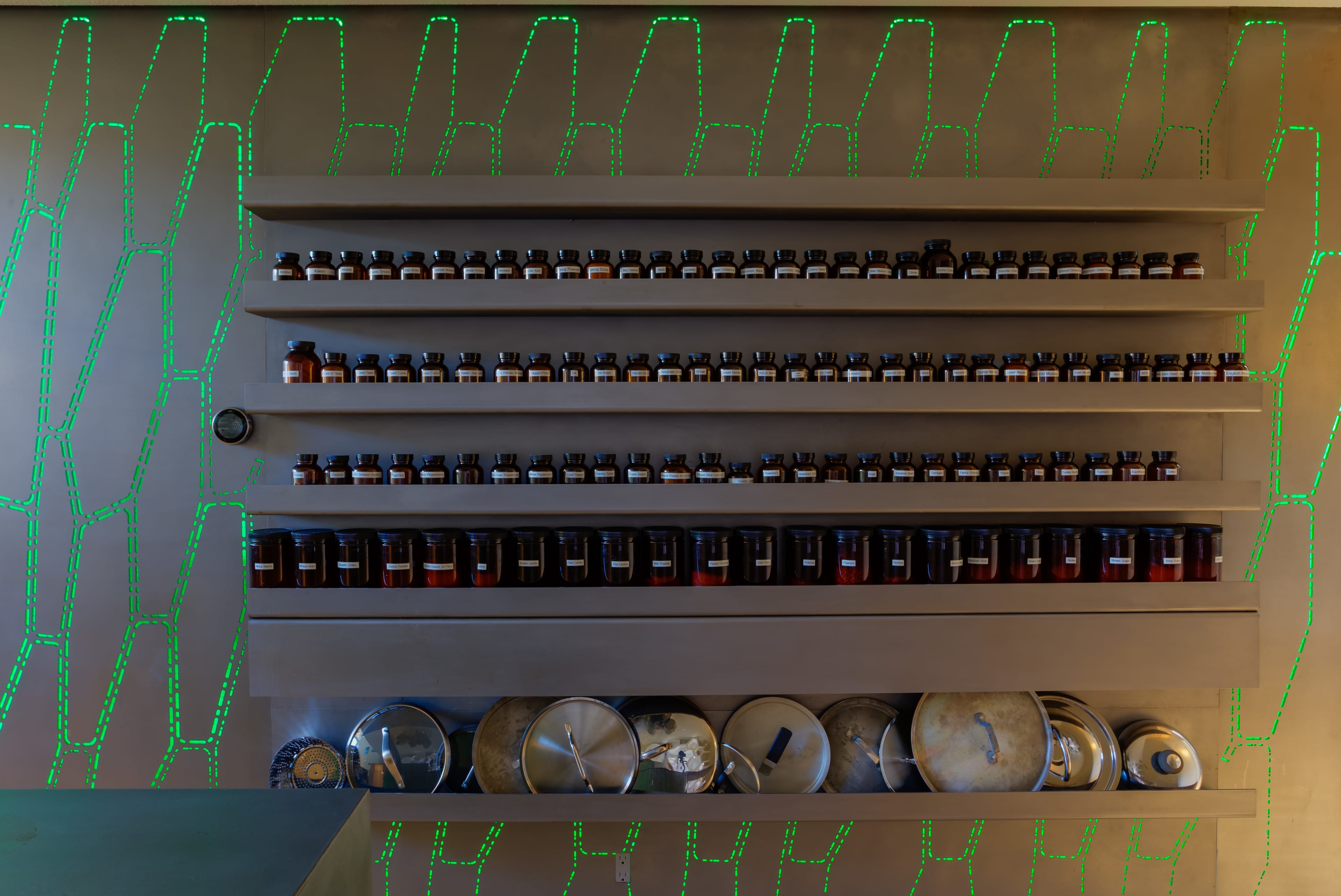 A wall-mounted shelf with neatly arranged jars and a row of metal lids below it, illuminated by a green pattern.