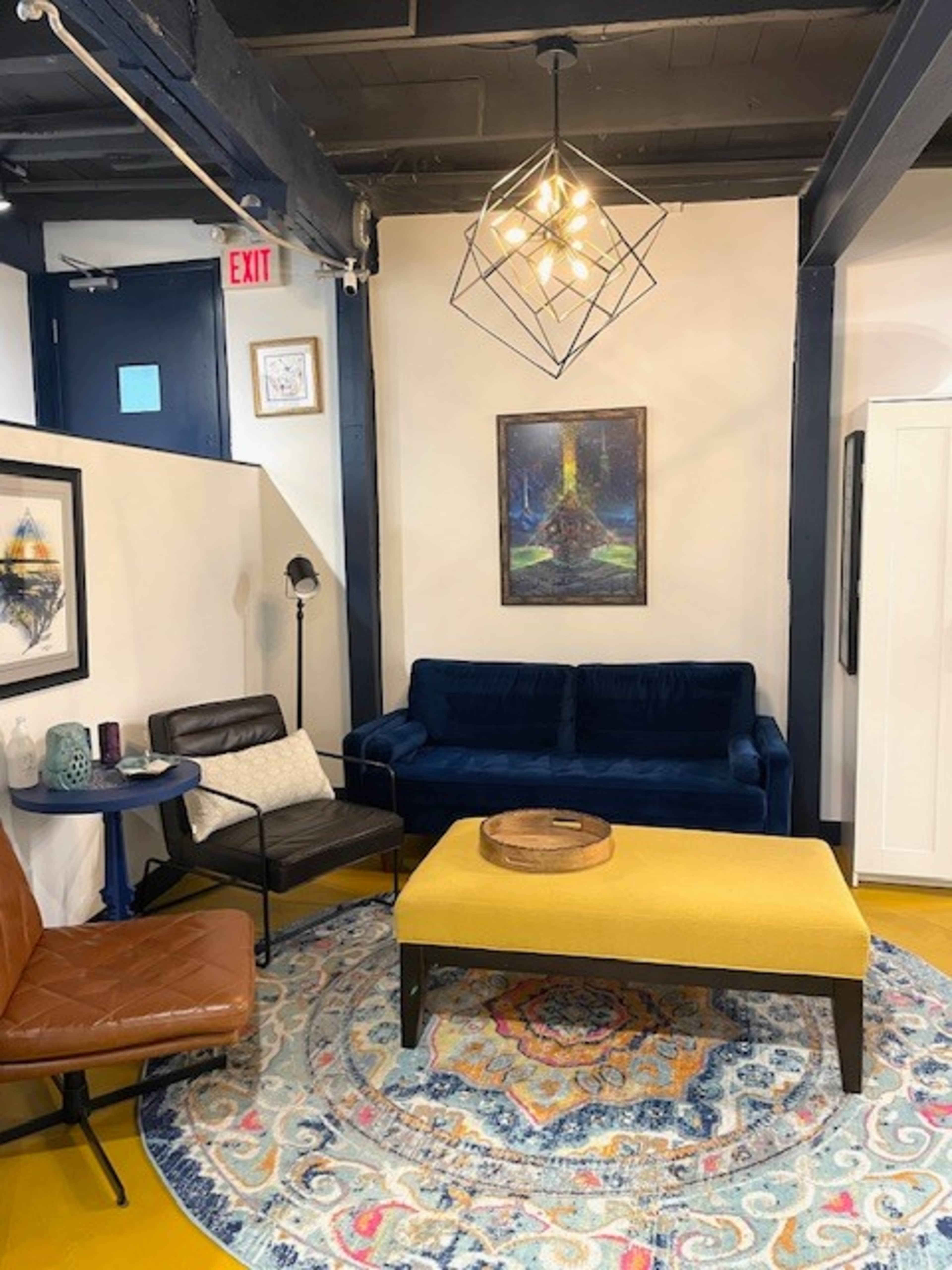 The image shows a cozy lounge area featuring a blue couch, a yellow coffee table, a patterned rug, and a piece of artwork on the wall.