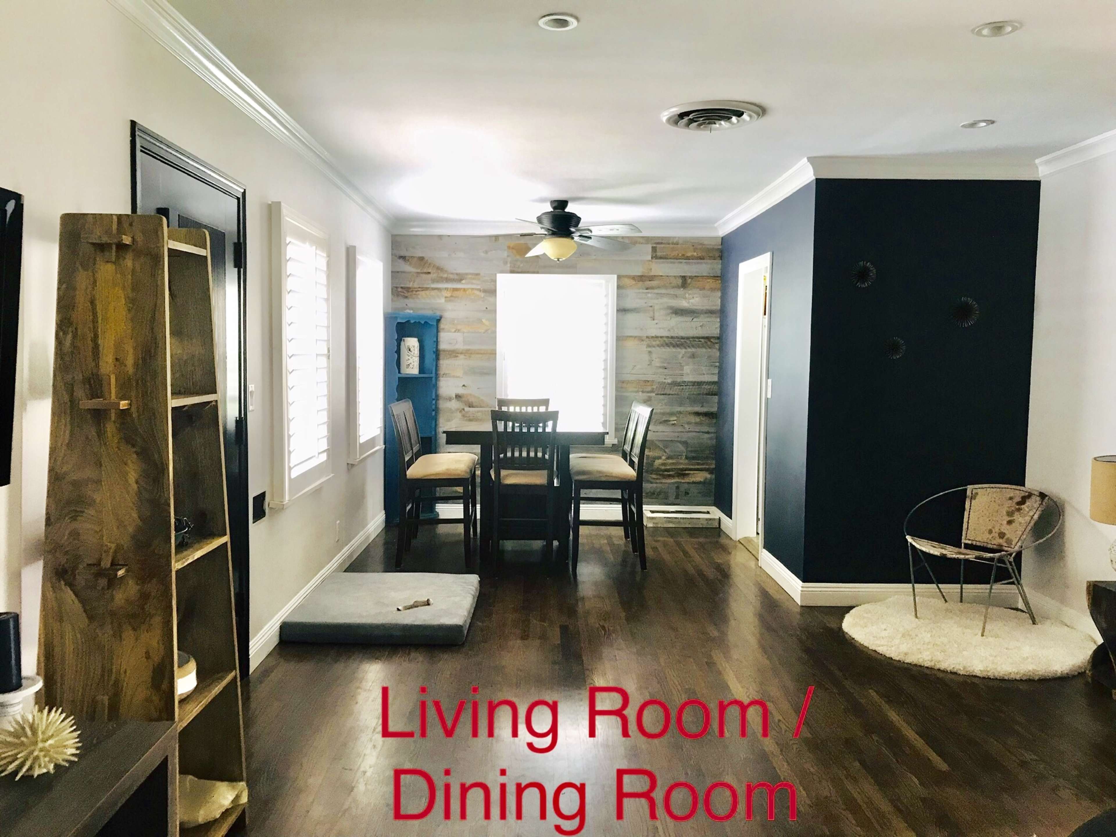 The image shows a living and dining room area featuring hardwood floors, a dining table with chairs, a bookcase, and a cozy chair.