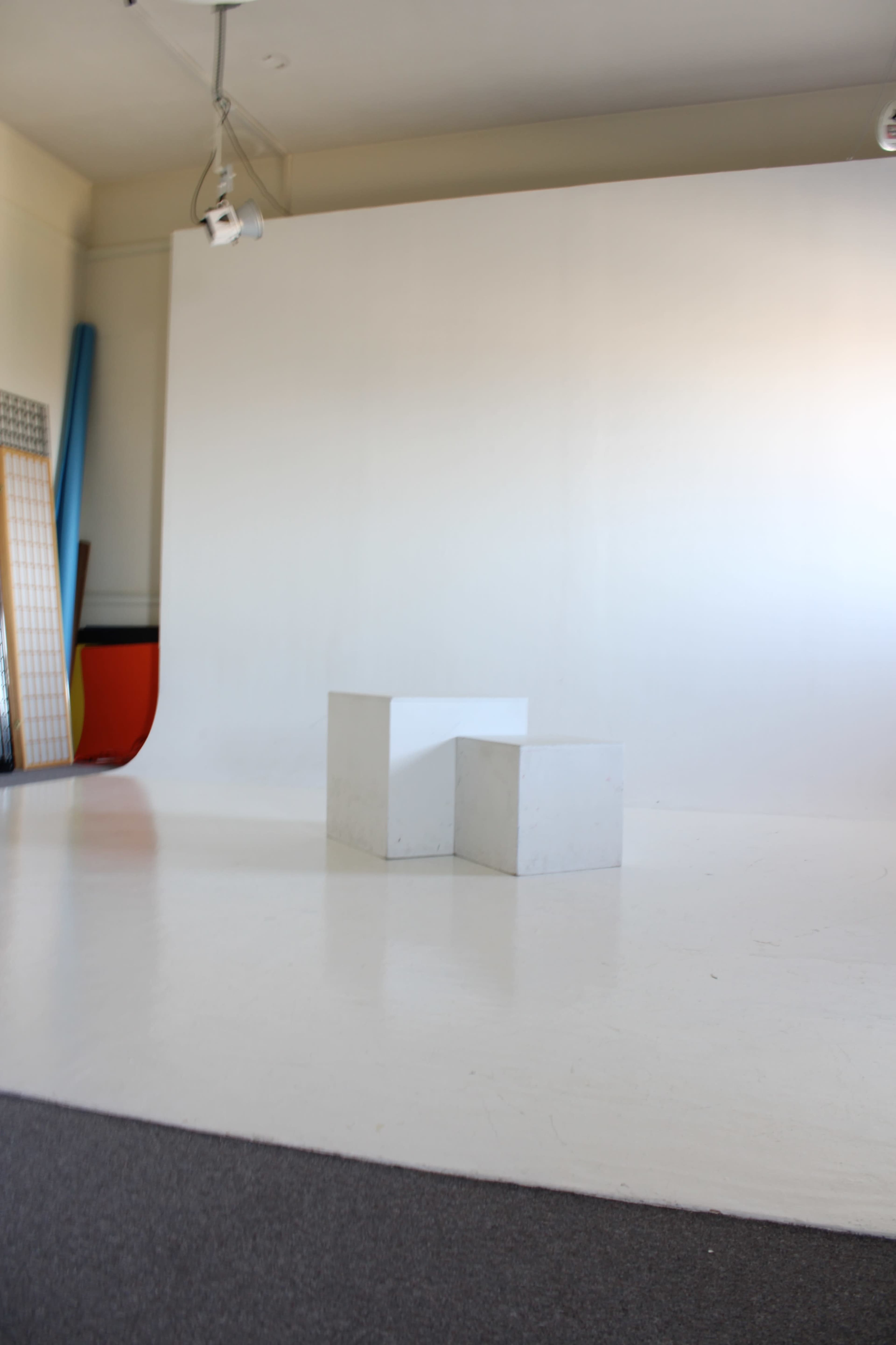 The image shows a minimalistic studio space with a white backdrop, a light-colored floor, and two white geometric blocks positioned in the center.