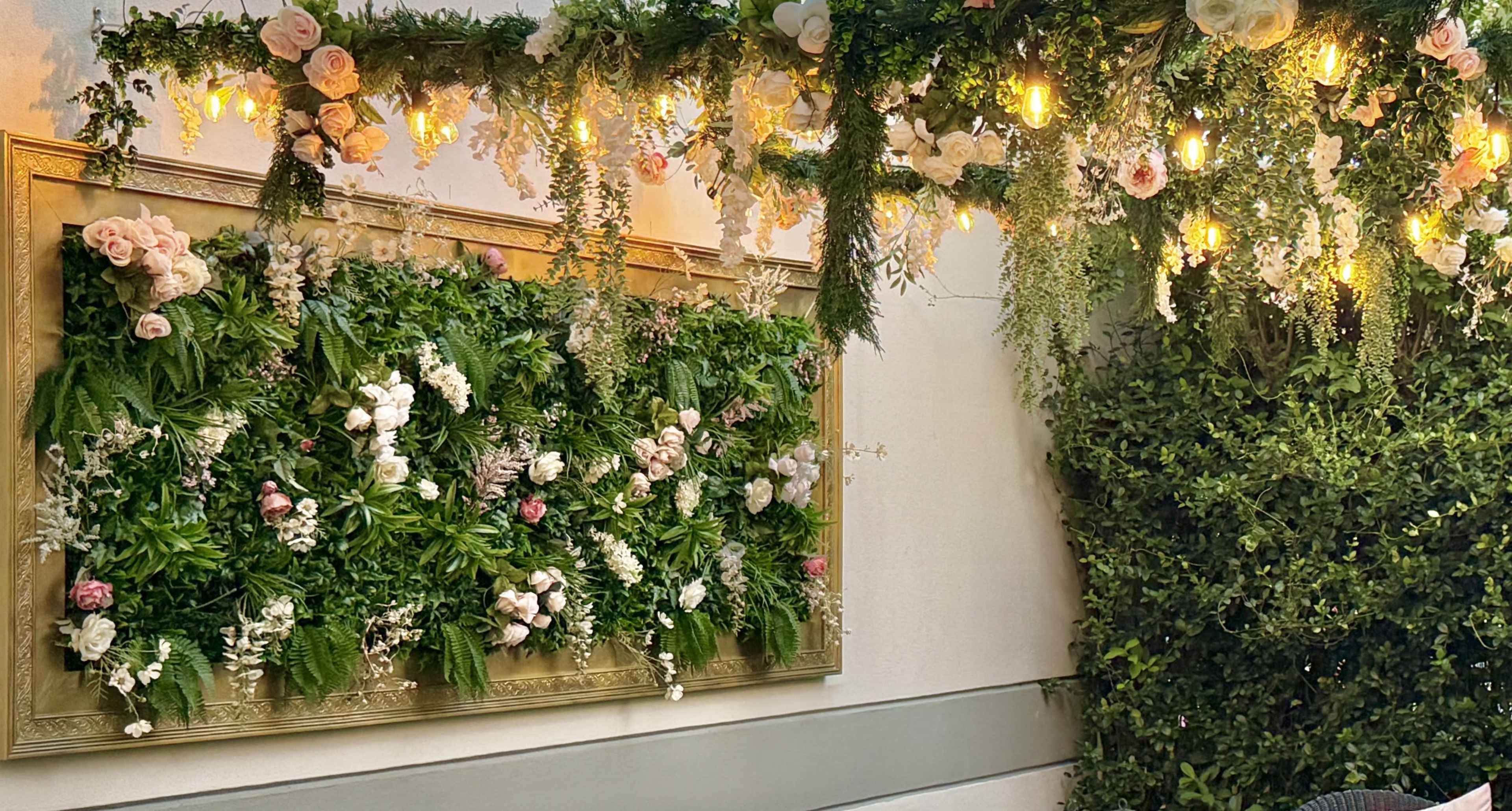 A floral wall display adorned with various greenery and flowers is illuminated by warm string lights, set against a white backdrop.
