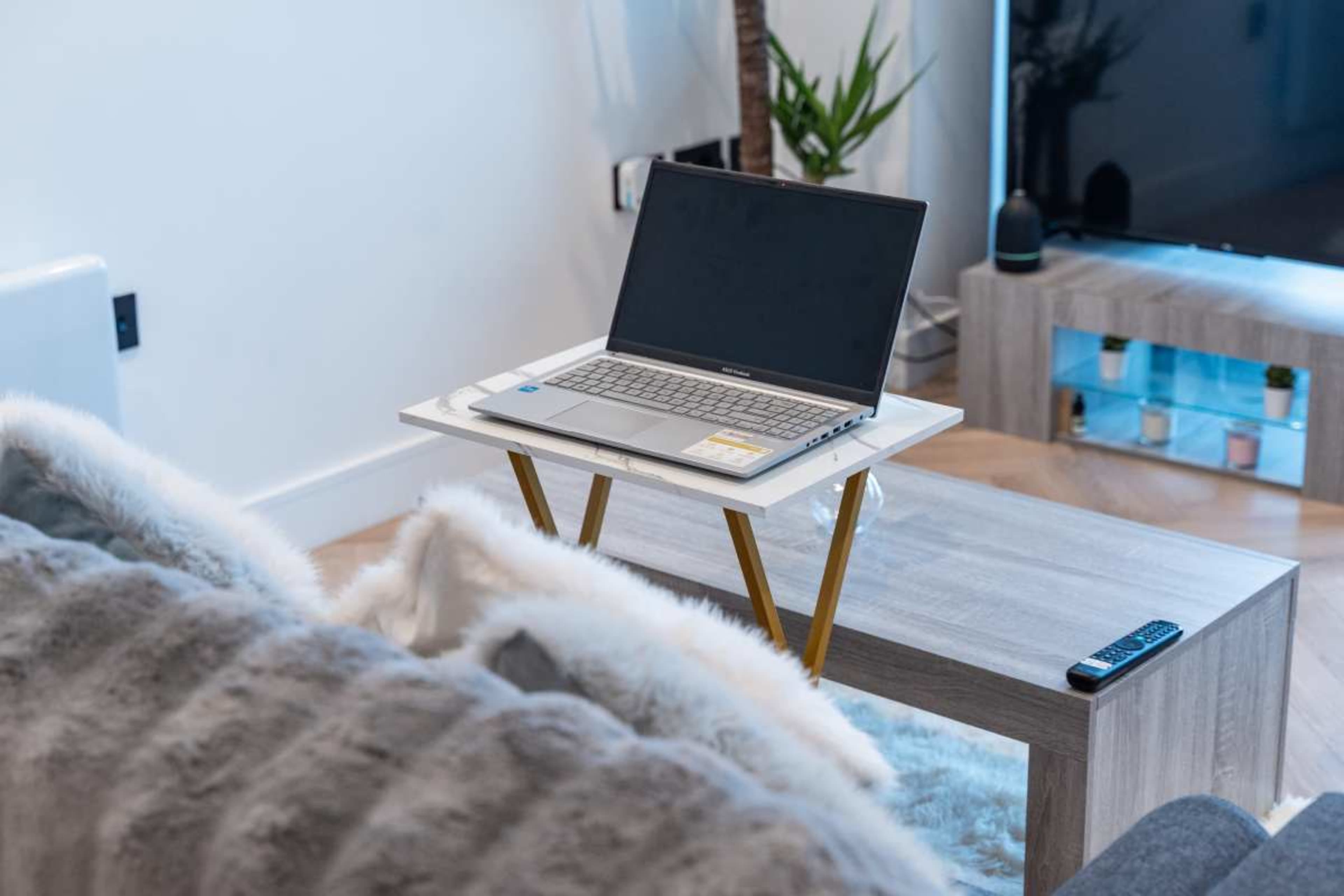 A laptop sits on a small table in a modern living room, surrounded by a couch and a television.
