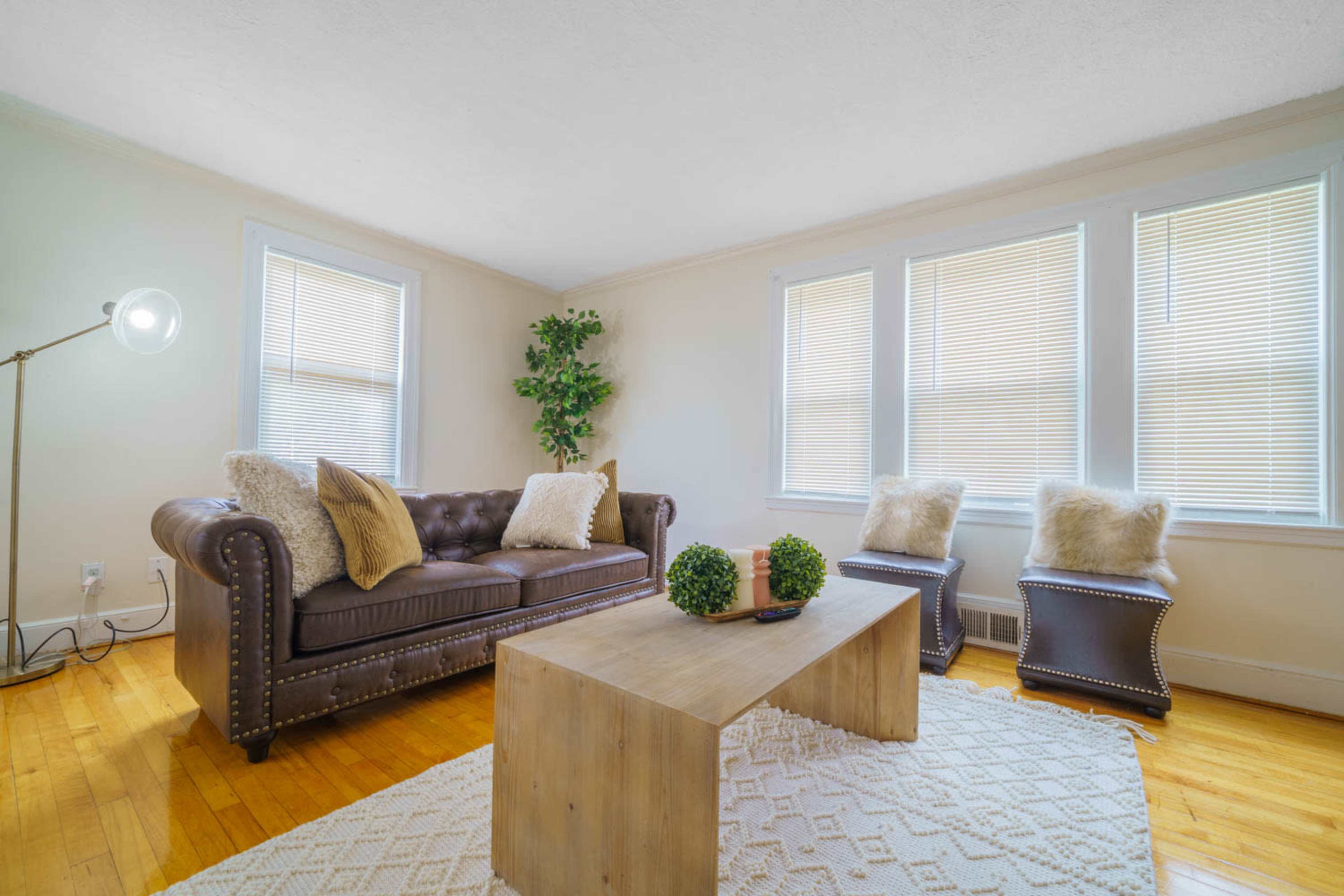 A cozy living room features a brown leather couch, a wooden coffee table, and two accent chairs positioned near large windows with blinds.