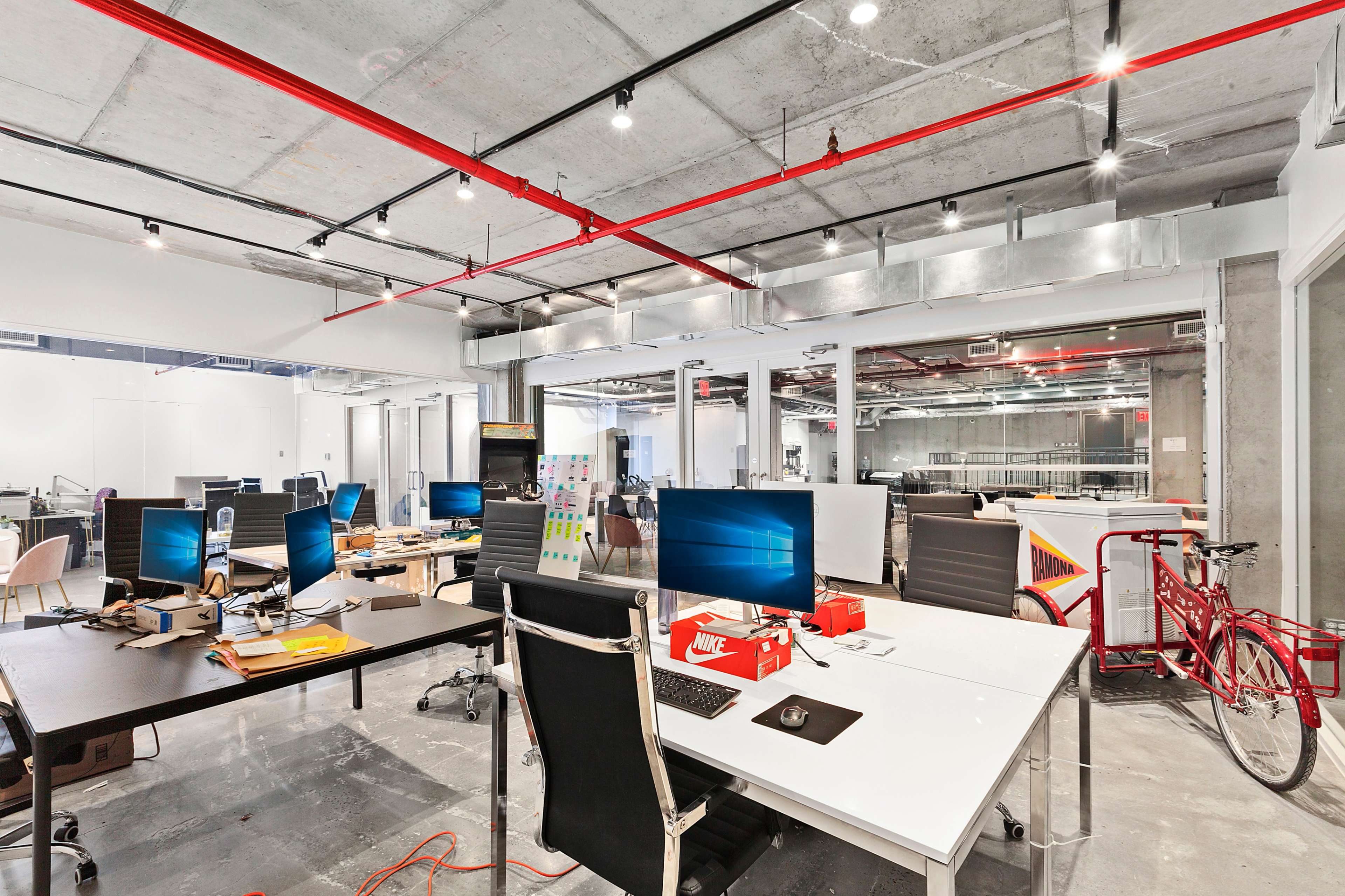 A modern office space with several desks, computers, and a bicycle, featuring exposed ceilings and a concrete floor.