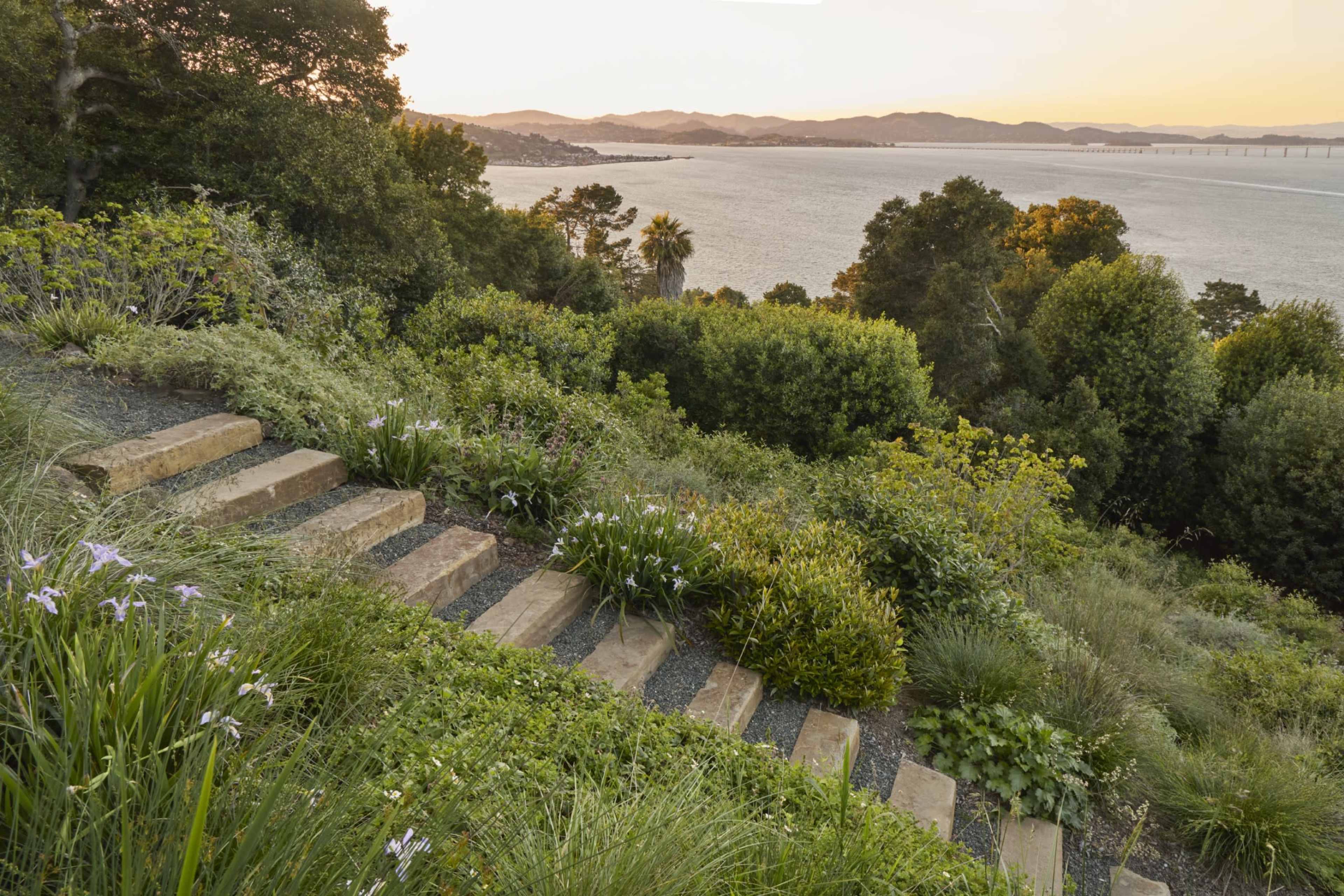 The image shows a staircase made of stone steps leading down through a lush garden toward a bay with hills in the background at sunset.