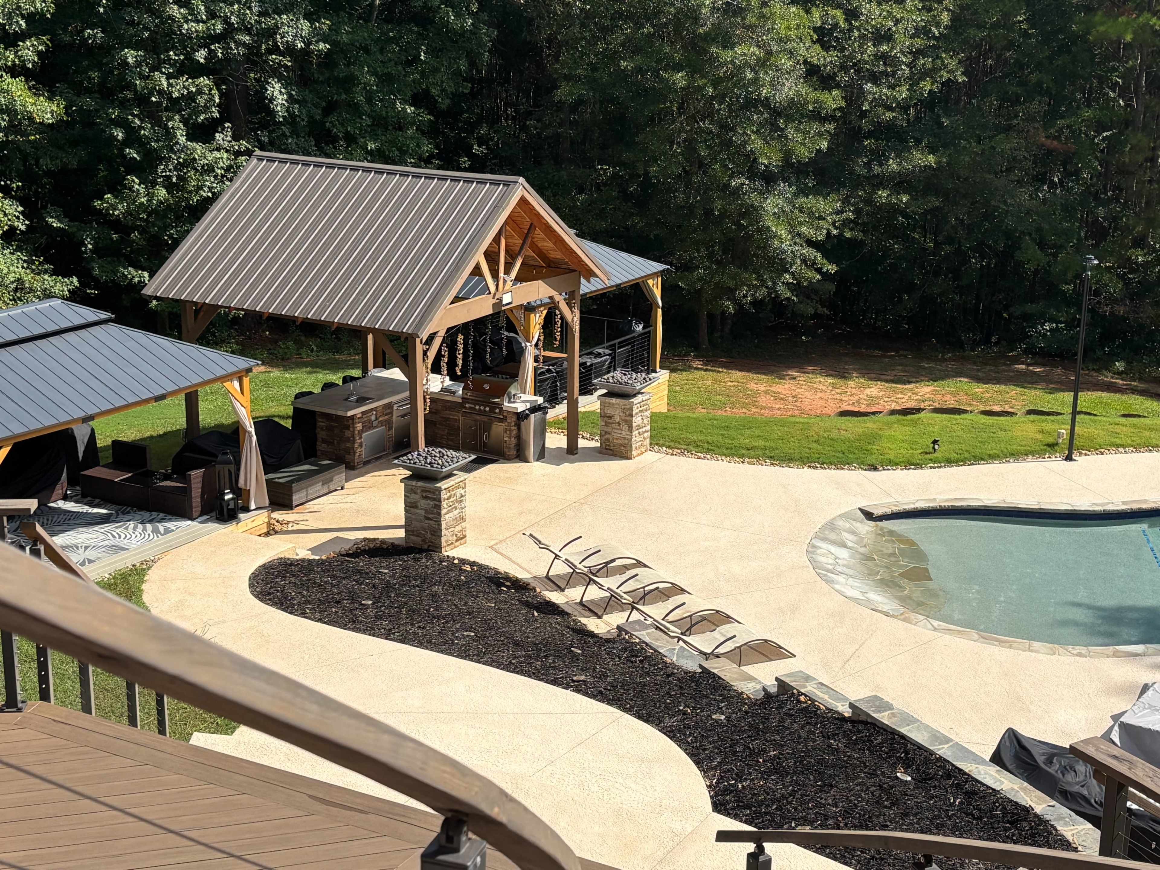 The image shows a backyard with a swimming pool, a stone patio, lounge chairs, and a covered outdoor kitchen area surrounded by trees.