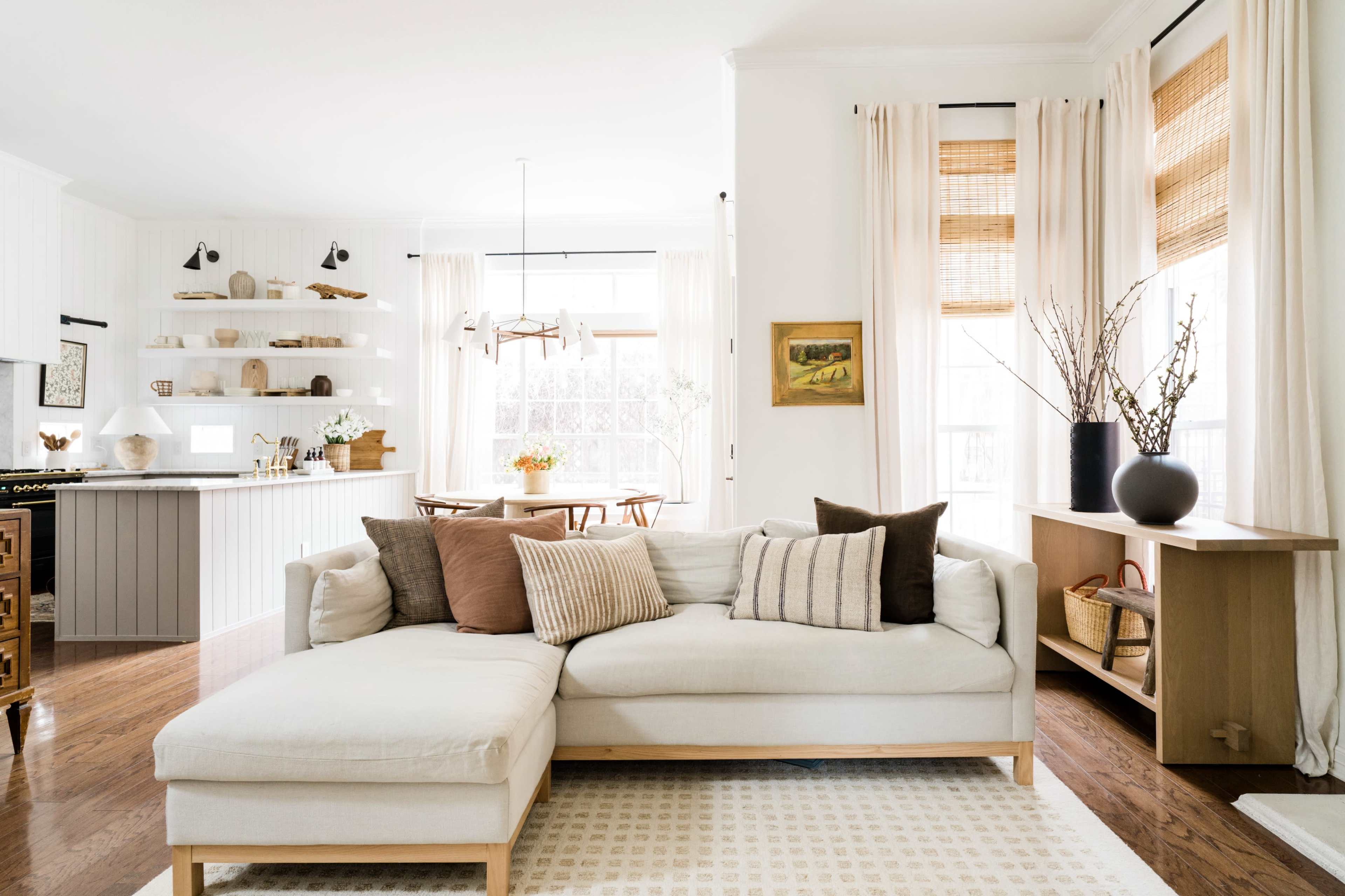 A bright living room features a light-colored sectional sofa with decorative cushions, a wooden side table, and large windows with woven shades.