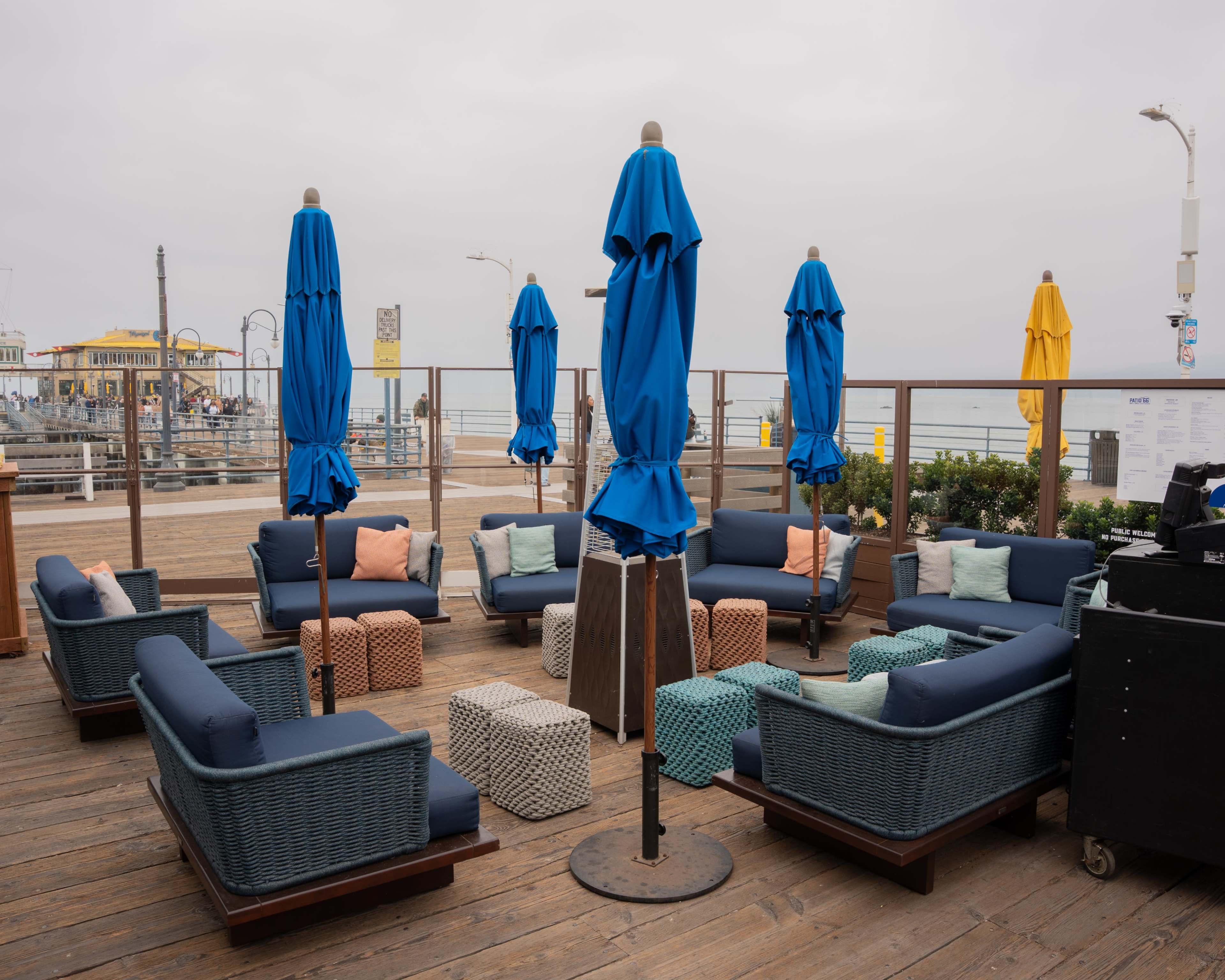A seaside patio features blue umbrellas and comfortable seating arranged around small tables on a wooden deck.
