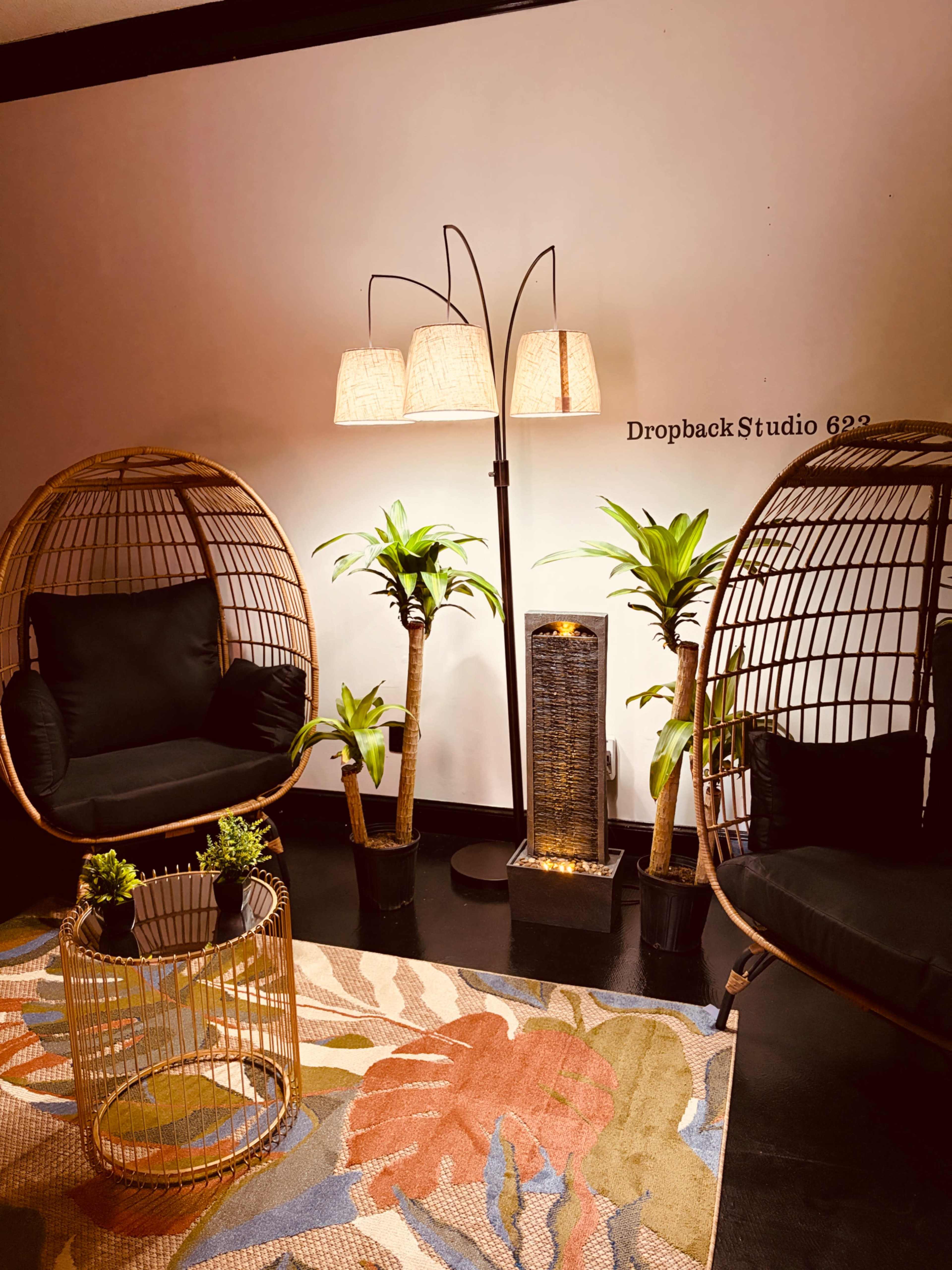 The image shows a cozy interior space with two rattan hanging chairs, a floor lamp with three light shades, a water feature, and potted plants on a patterned rug.