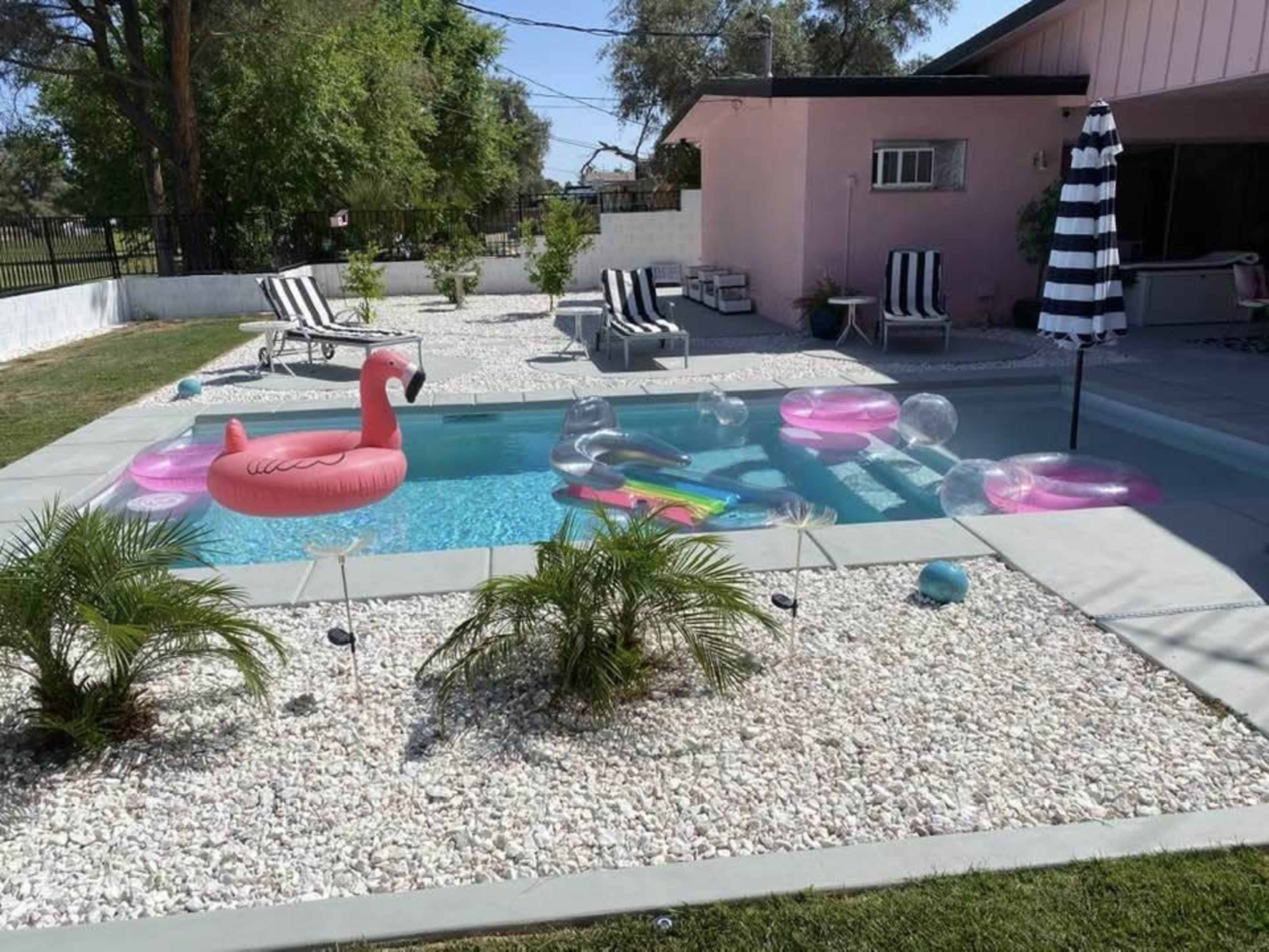 A swimming pool surrounded by gravel and lounge chairs features colorful inflatable floats, including a flamingo and a striped umbrella.
