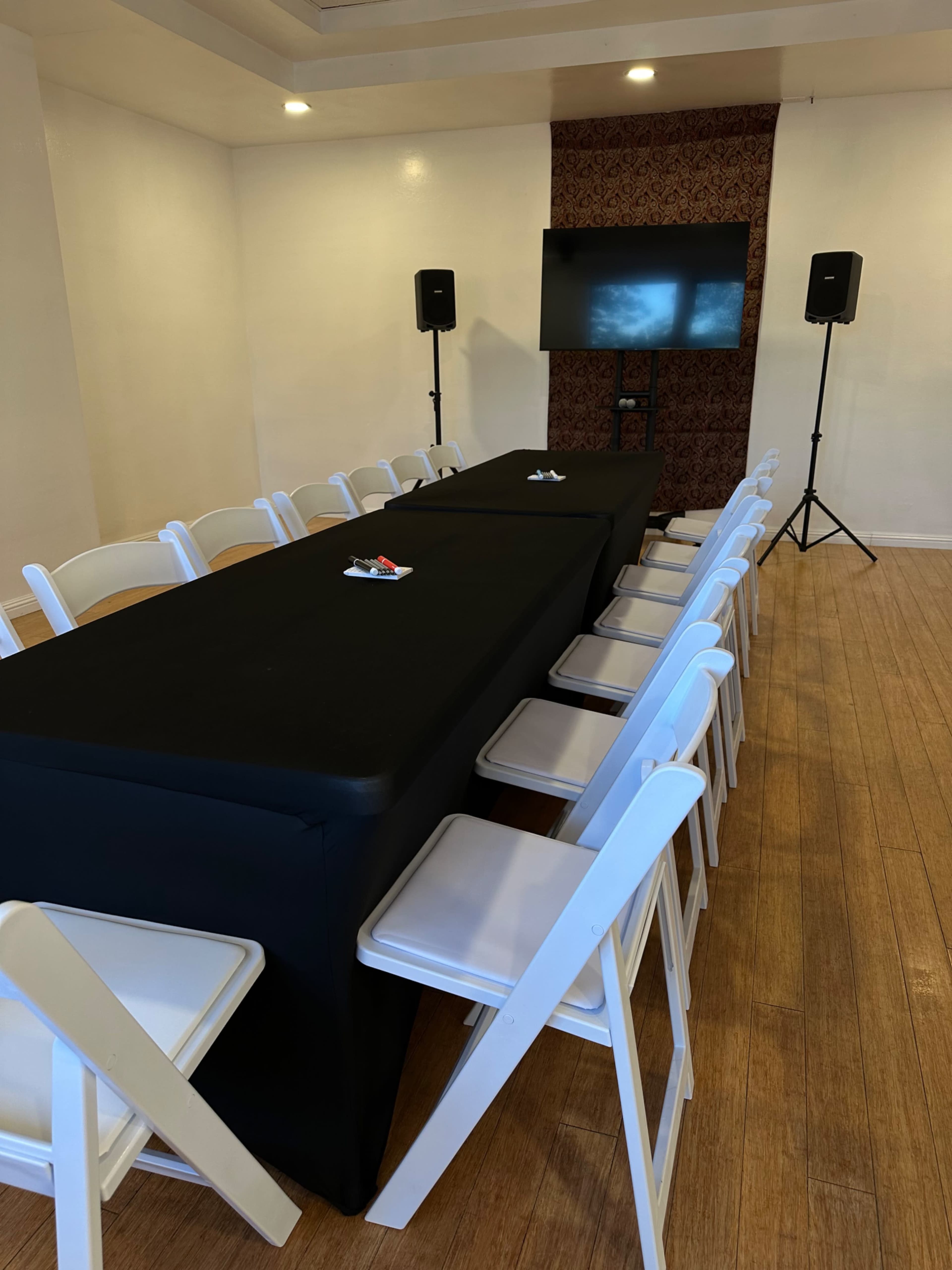 A long black table is set with white chairs in a brightly lit room, featuring a television on the wall and speakers positioned nearby.