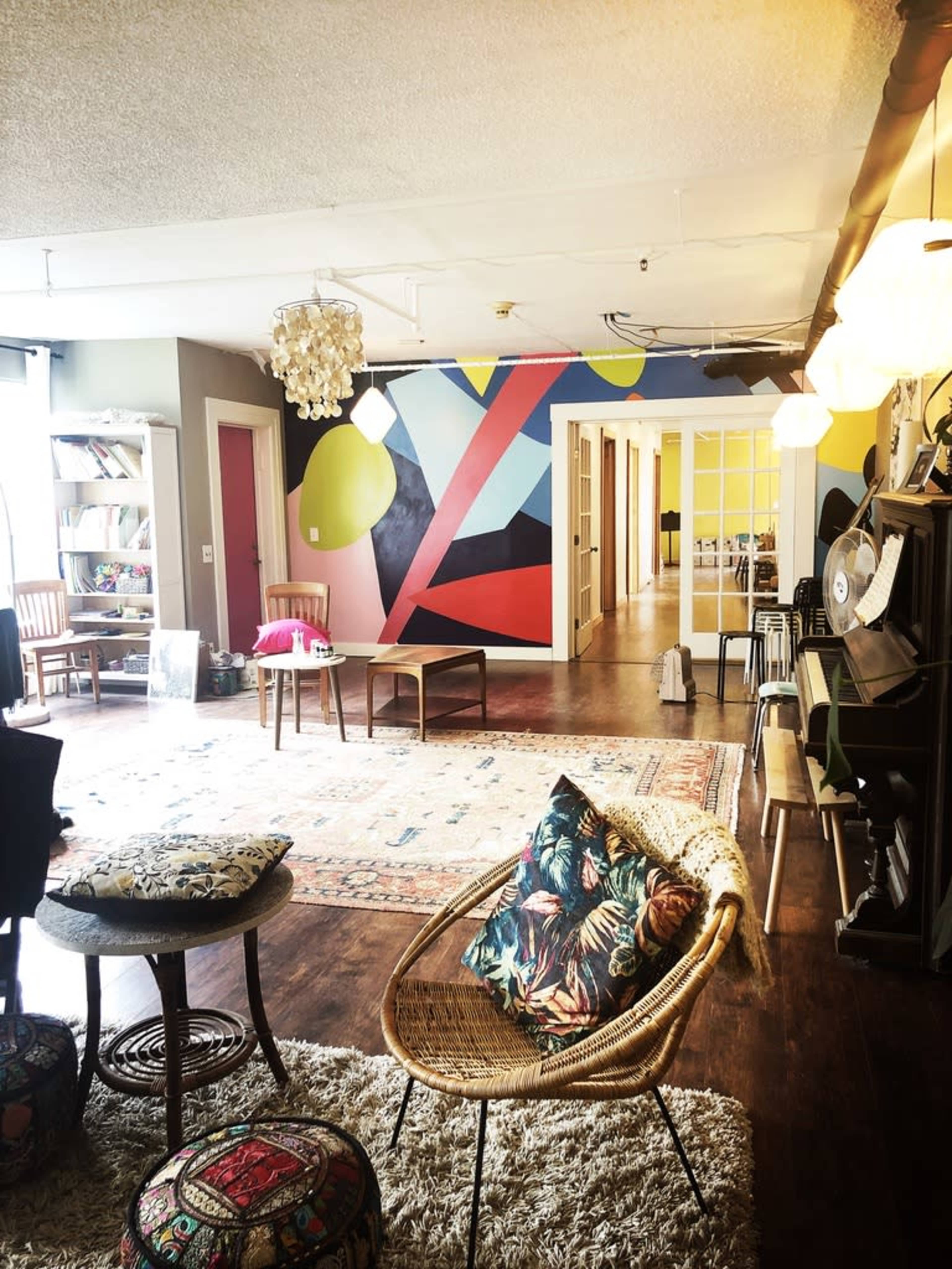 A spacious, well-lit room with a colorful mural on one wall, featuring various shapes, and furnished with eclectic seating arrangements including a rattan chair and patterned cushions.