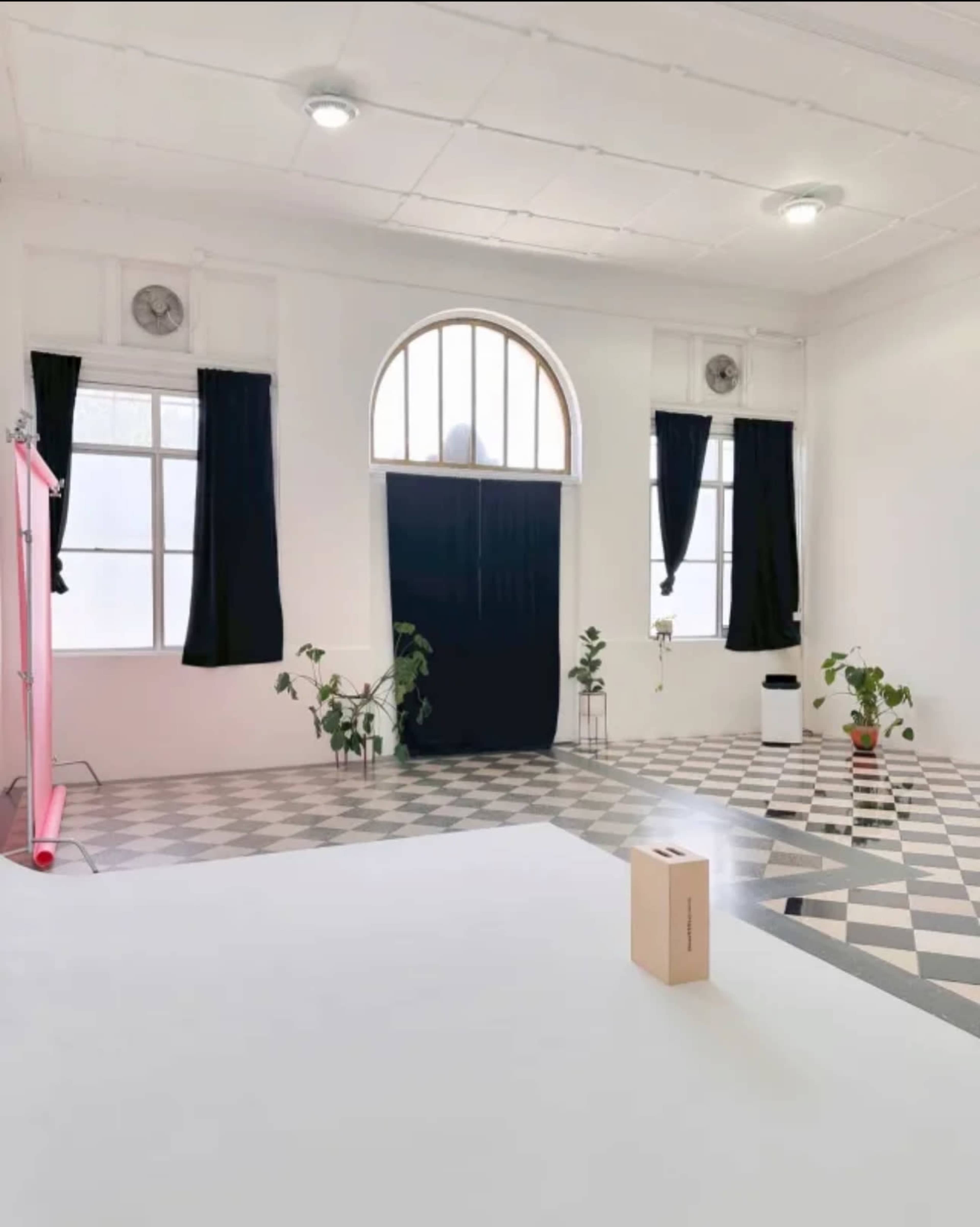 The image shows a spacious, minimally decorated room with black curtains, potted plants, and a box on the floor, featuring patterned tiles and large windows.