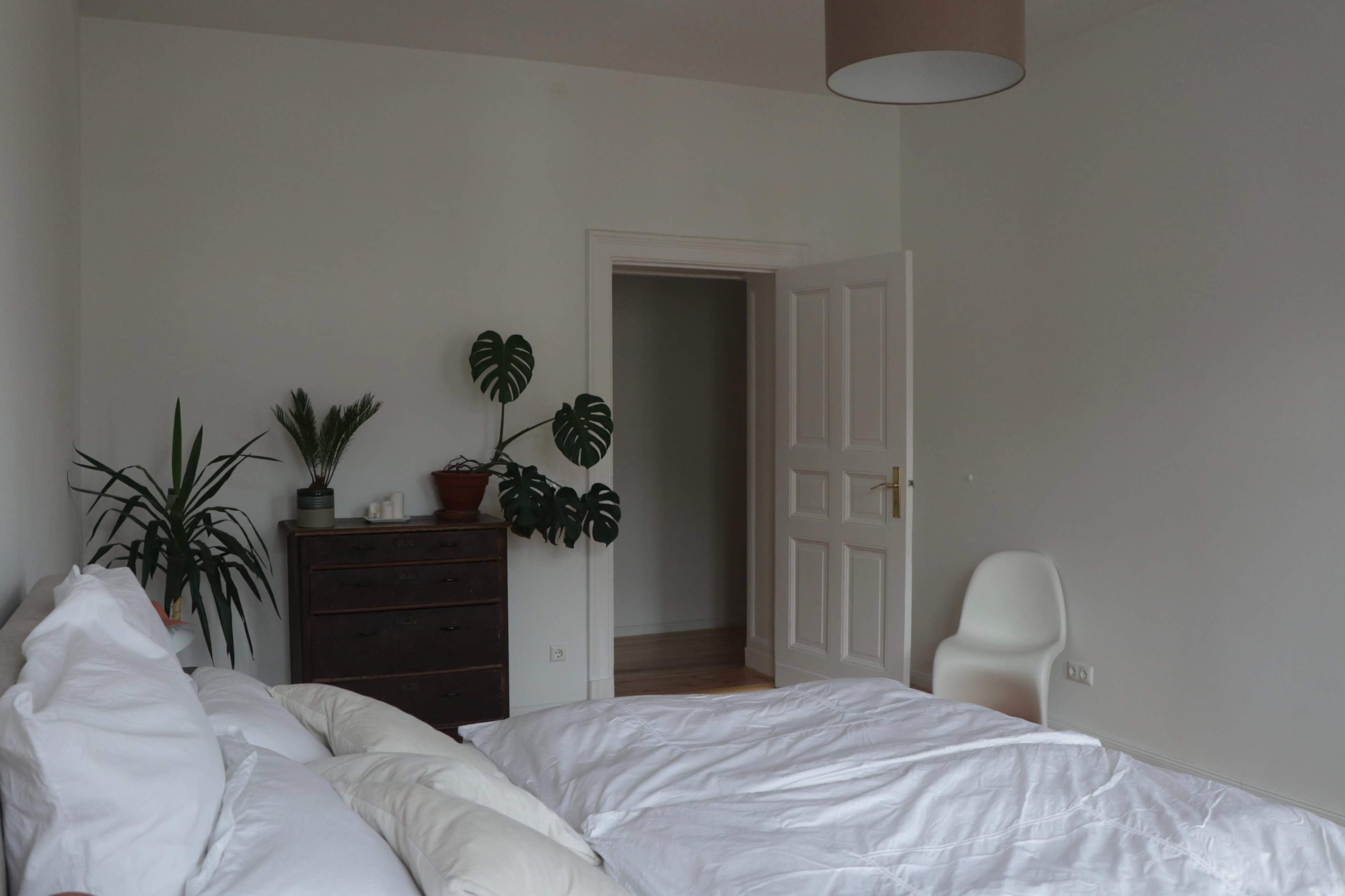 A minimalistic bedroom features a bed with white linens, a dark wooden dresser, a green plant on top, and an open door leading to another room.