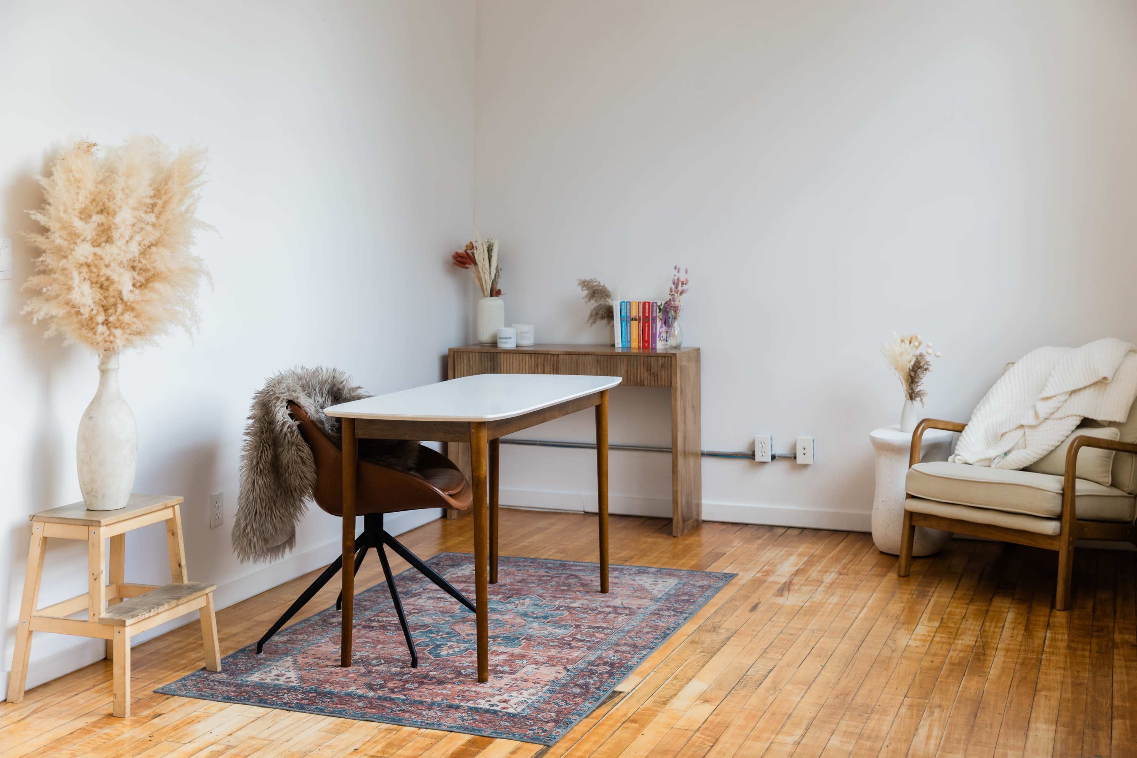 The image shows a minimalist home office setup featuring a wooden desk with a chair, a decorative rug, a small bookshelf, and a cozy seating area.