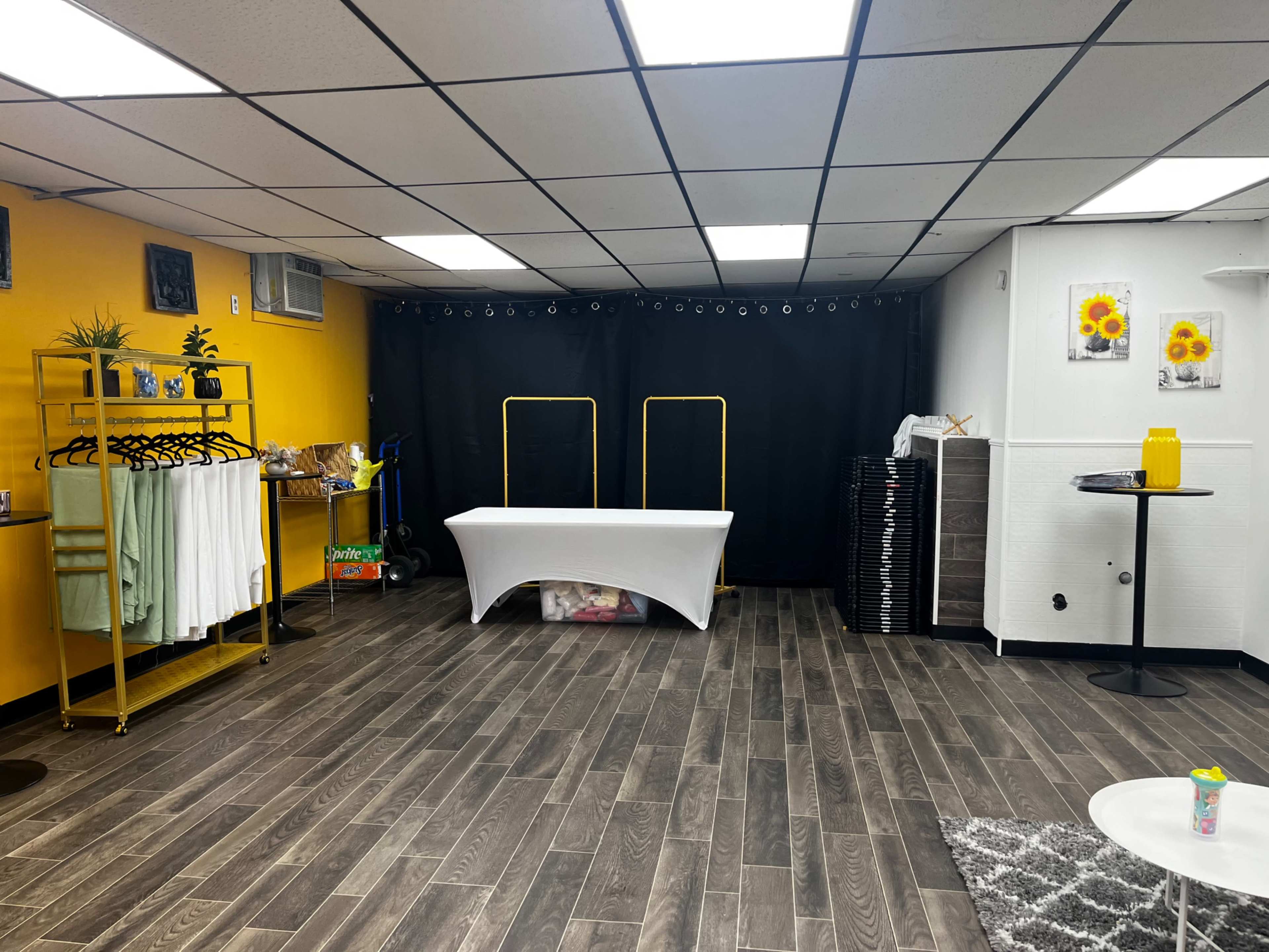 The image shows a room with gray wooden floors, yellow walls, and a white table in the center, surrounded by various equipment and decorative elements, including a clothing rack and black curtains.