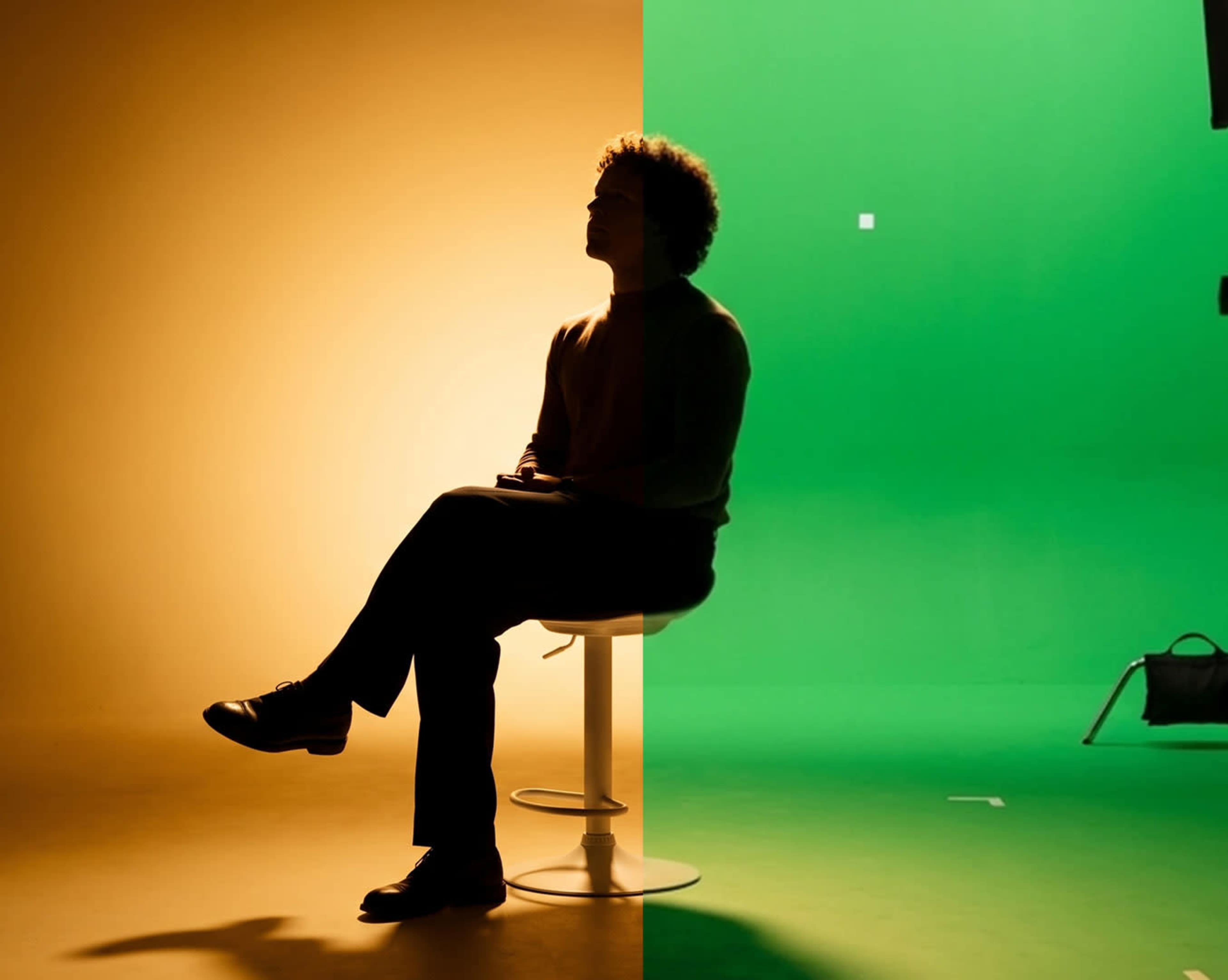 A person with curly hair sits on a stool, half of the background illuminated in orange and the other half in green.