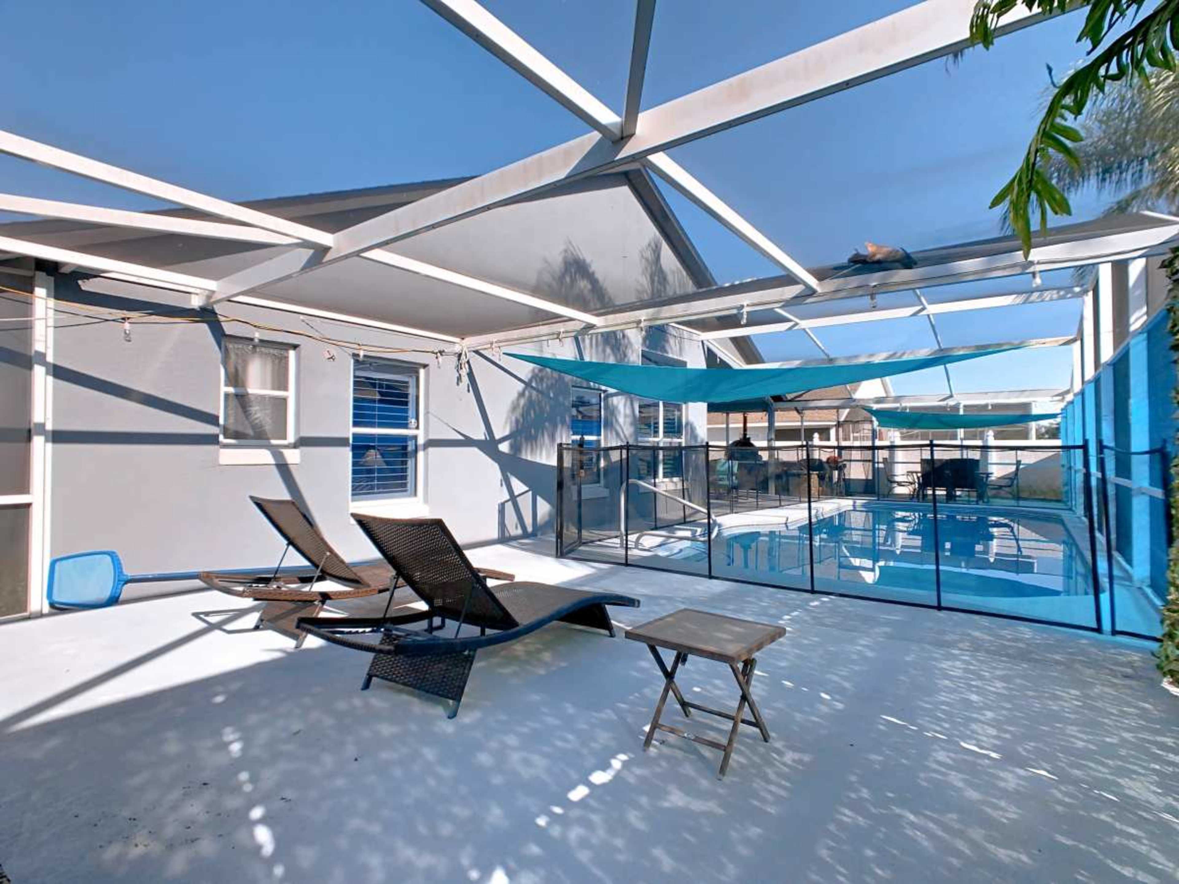 The image shows a backyard pool area with lounge chairs, a table, and a fenced-in pool under a blue sky.
