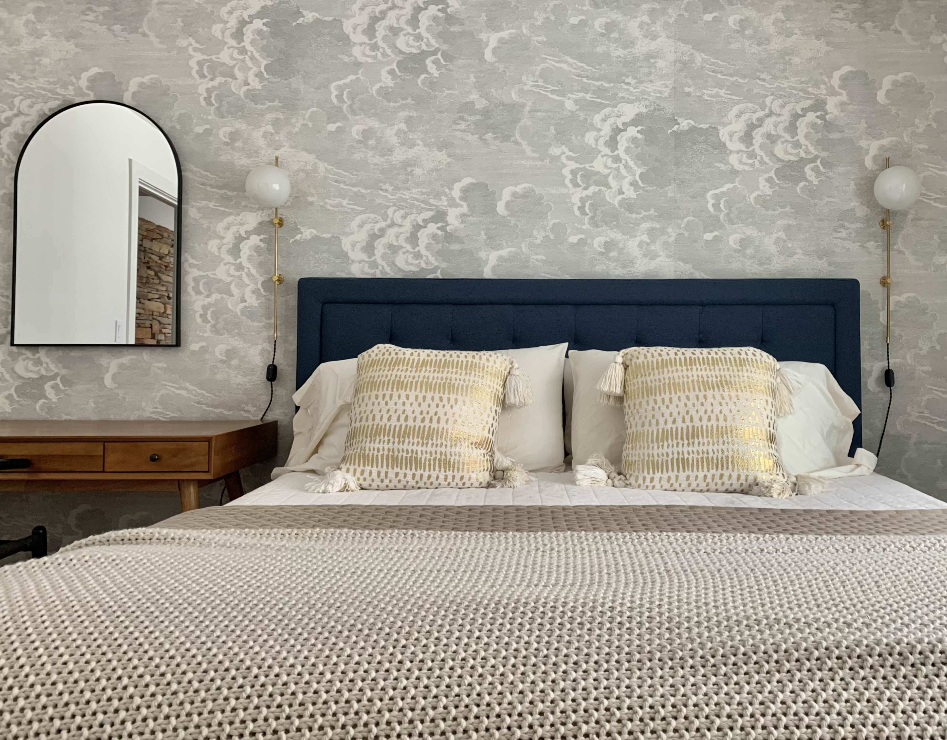 A neatly made bed with blue upholstered headboard and decorative pillows is positioned against a cloud-patterned wall, accompanied by a wooden bedside table and a round mirror.