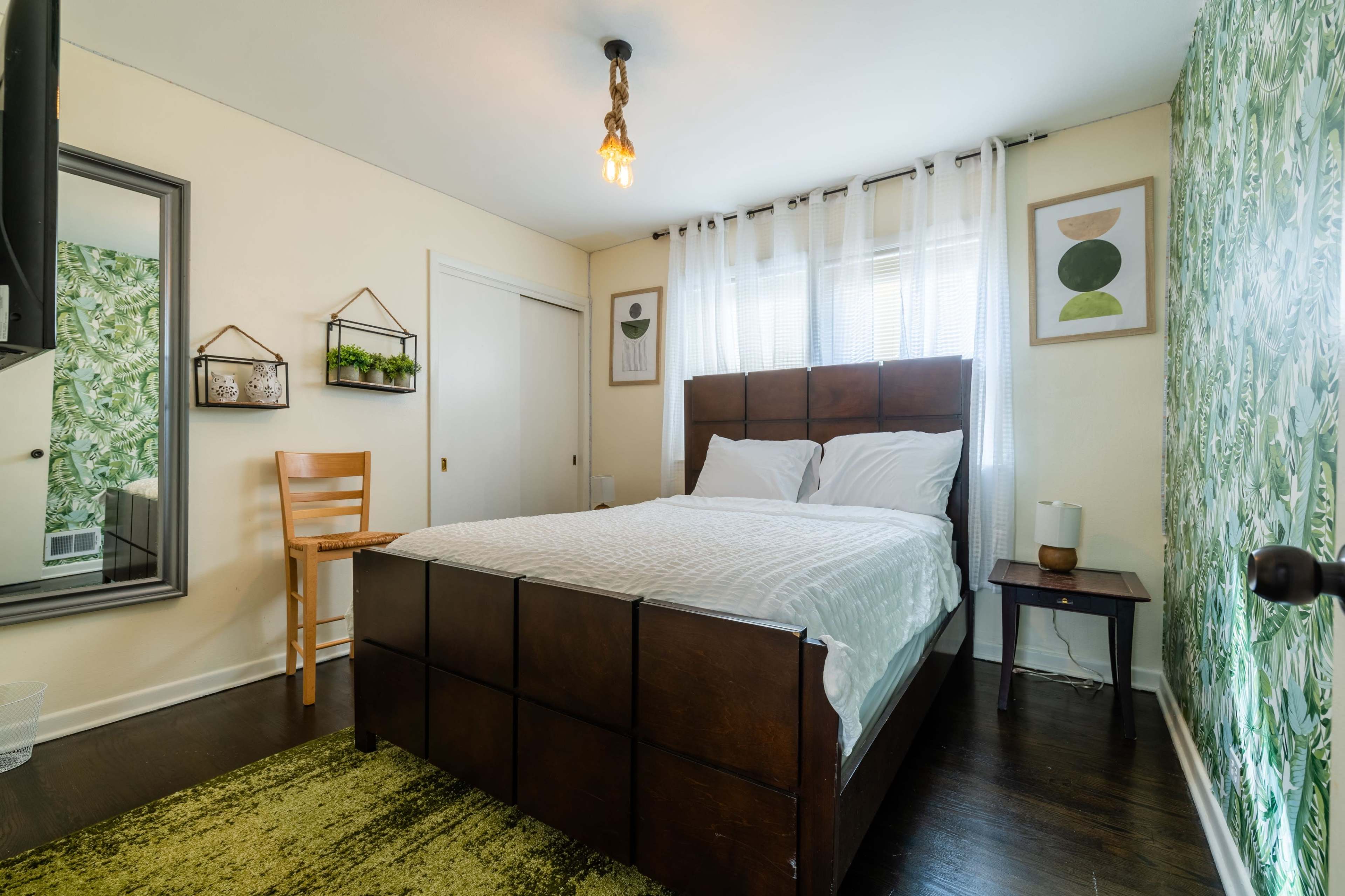 The image shows a cozy bedroom featuring a wooden bed, a chair, a nightstand, and decorative green wall art.