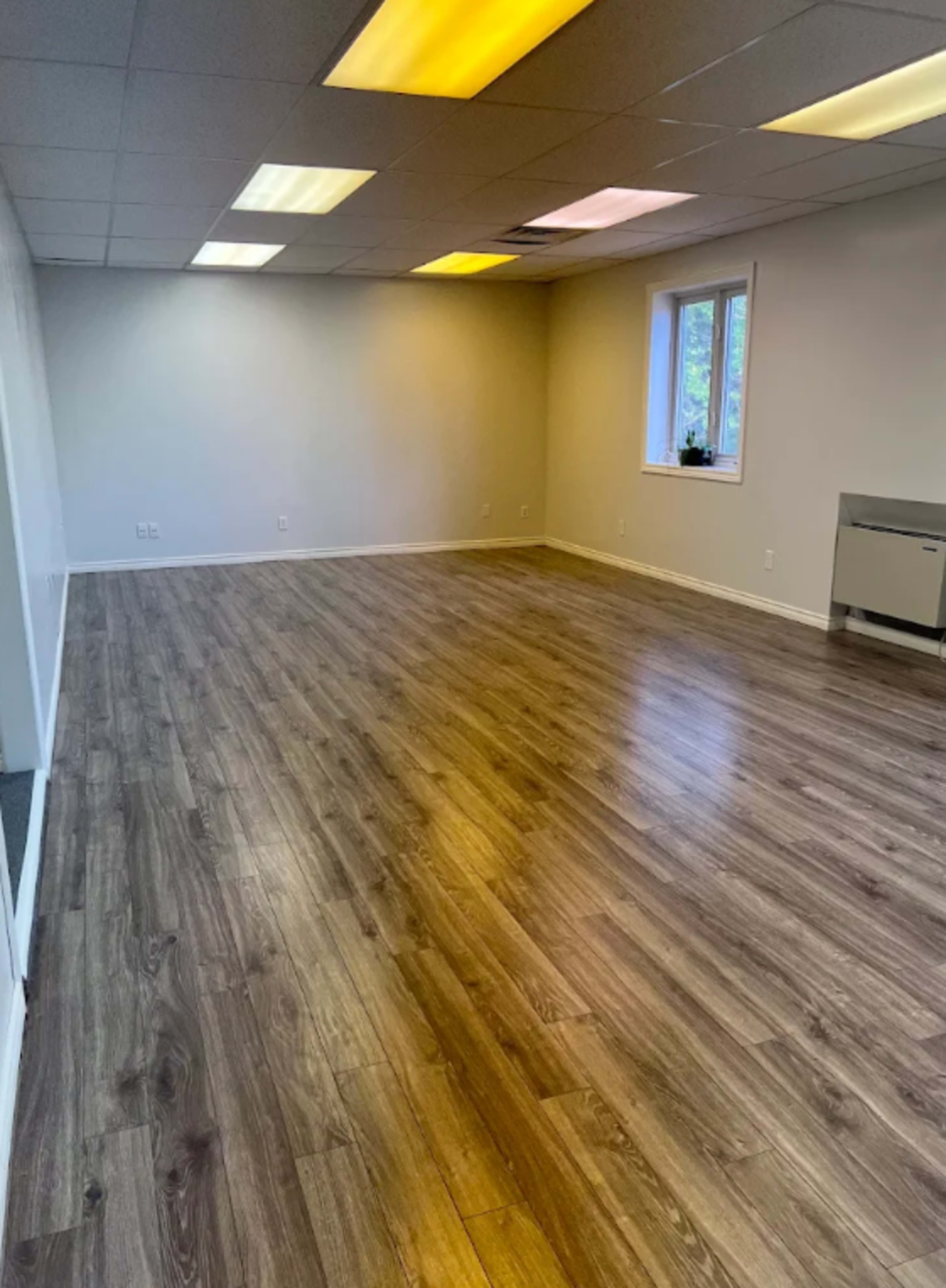 The image shows a spacious, empty room with wood laminate flooring and overhead fluorescent lighting.