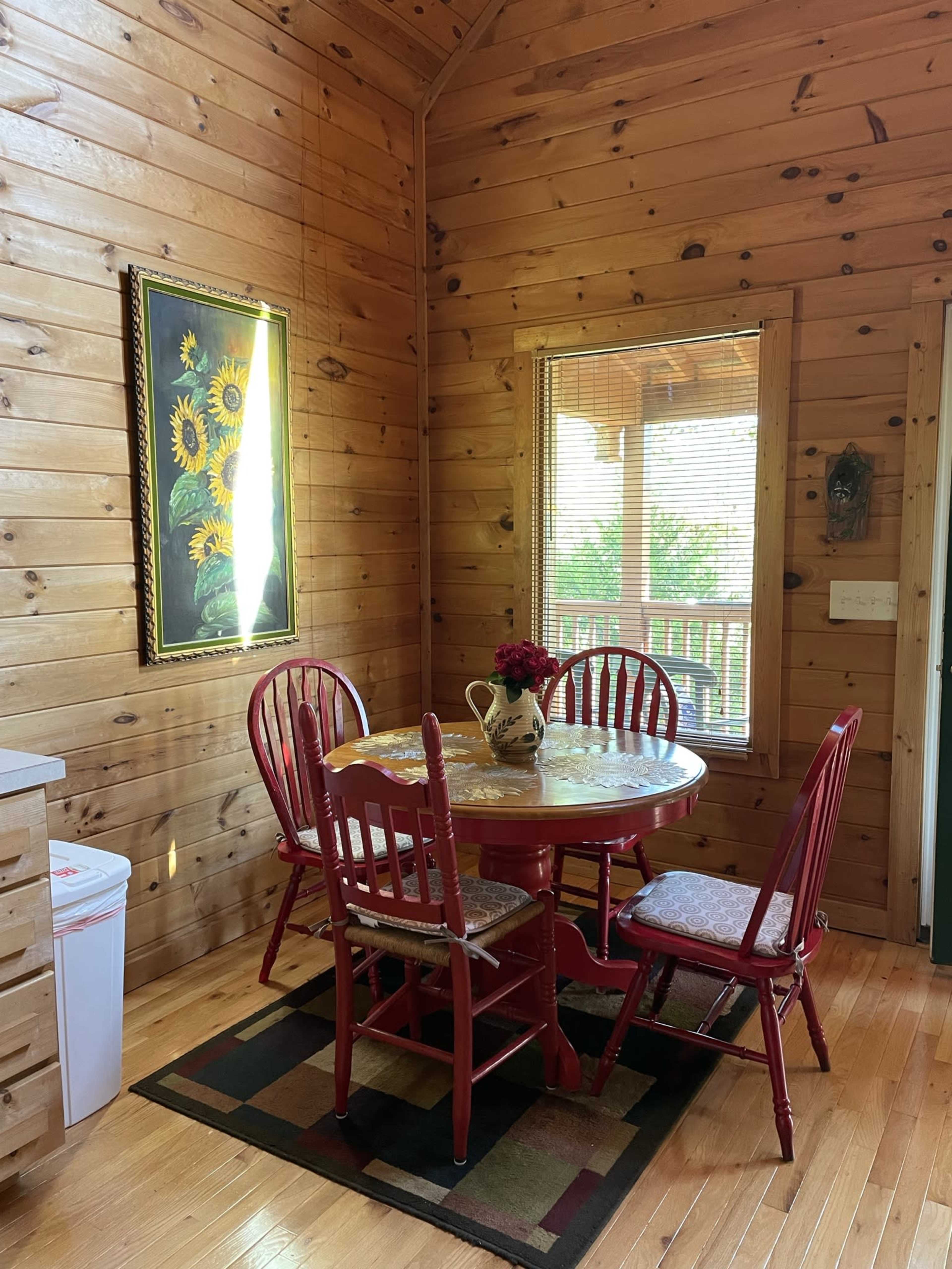 A wooden dining area features a round table surrounded by red chairs, with a vase of flowers and a sunflower painting on the wall.
