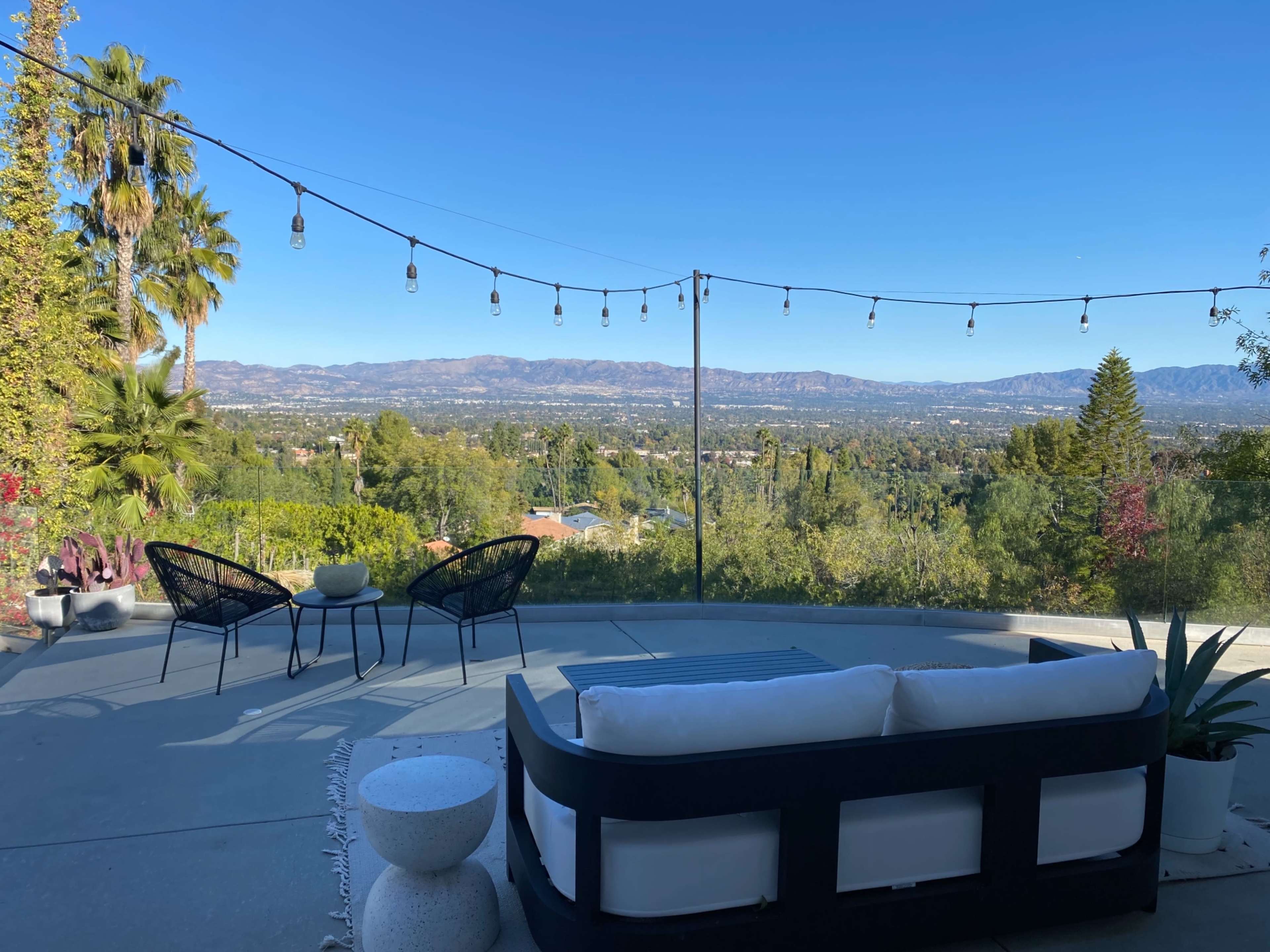 A patio with modern seating overlooks a valley and mountains under a clear blue sky, illuminated by string lights.
