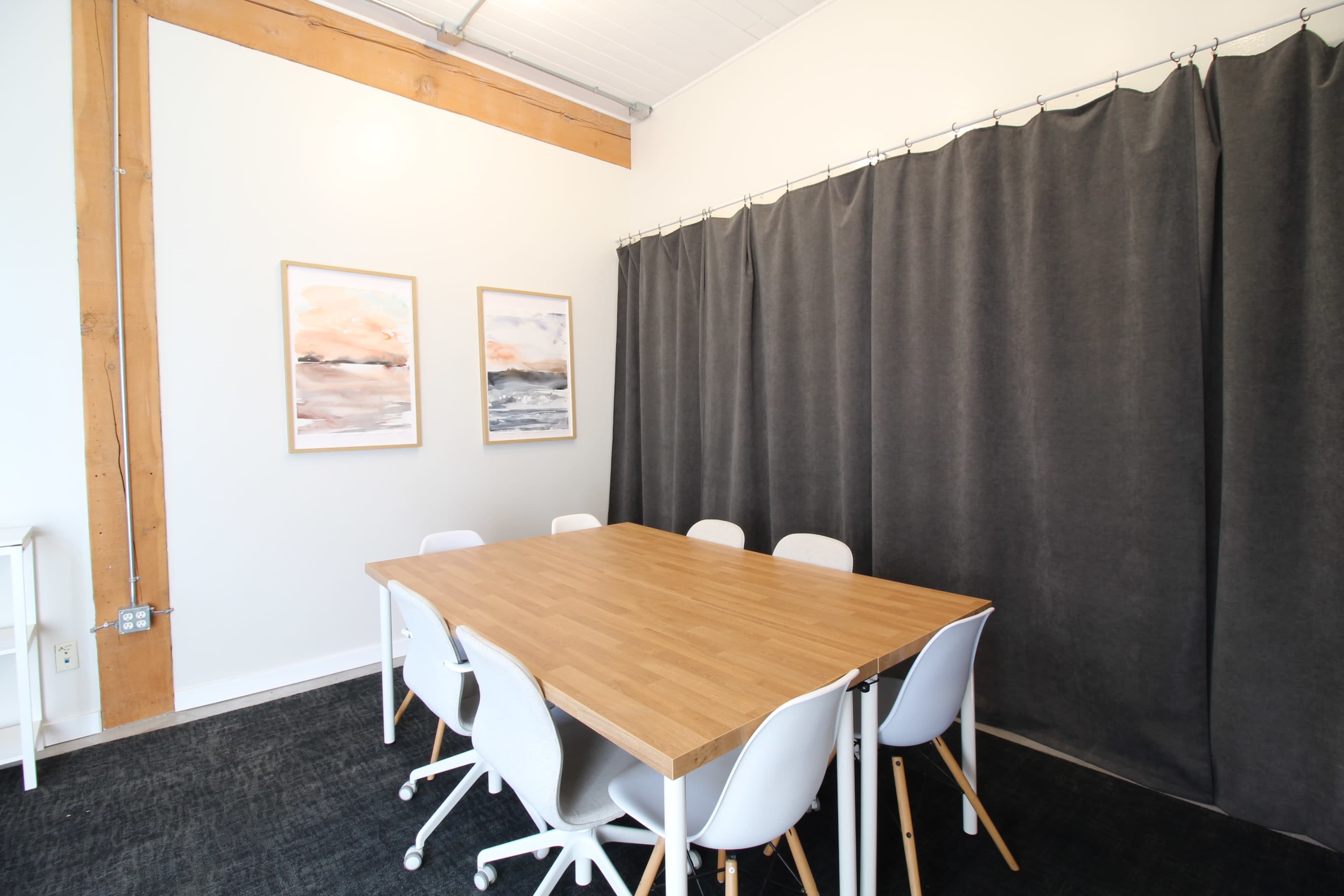 The image shows a meeting room with a wooden table surrounded by six white chairs and two framed artworks on the wall, adjacent to a gray curtain.
