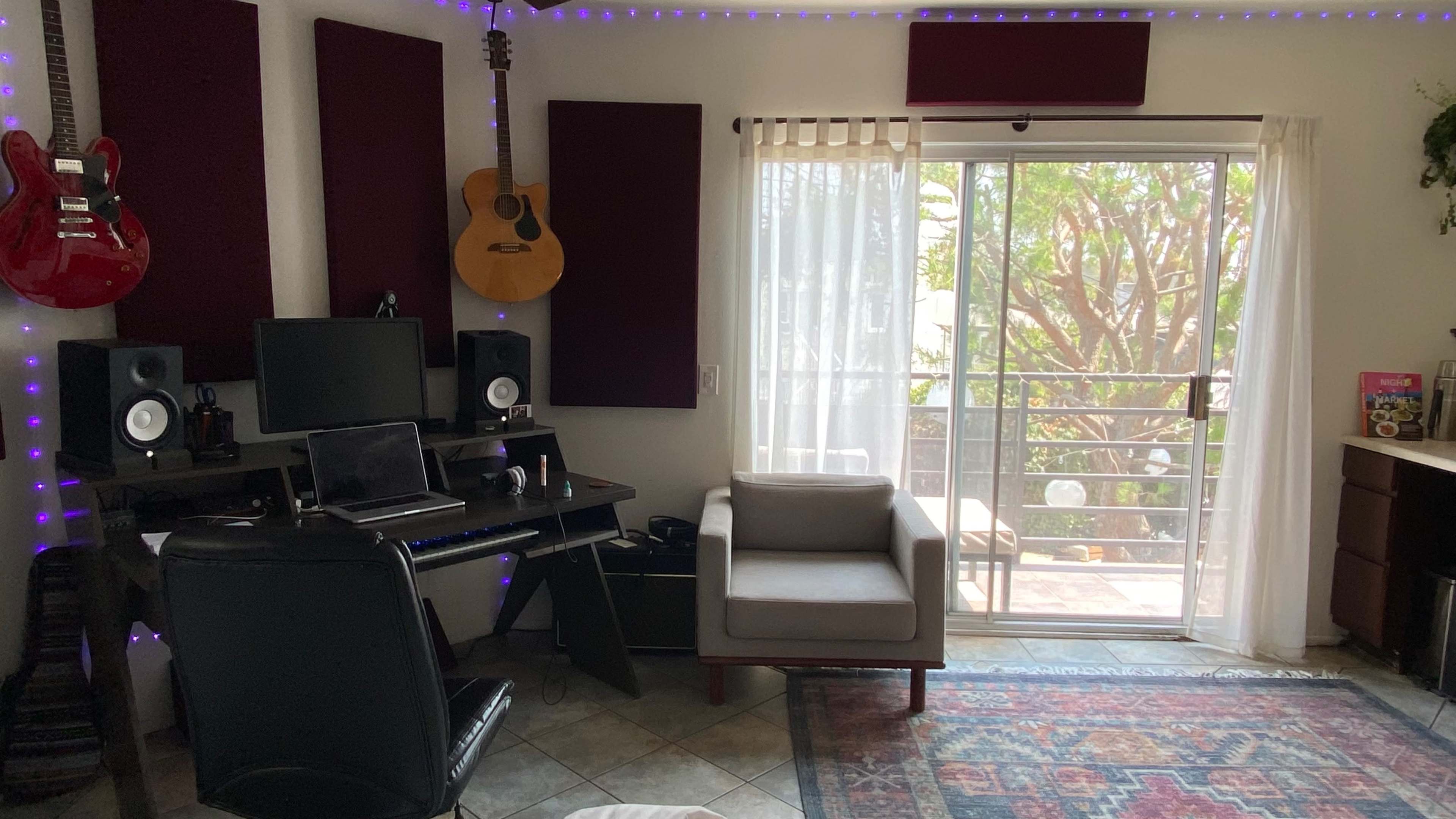 A home studio features guitars on the wall, a desk with a computer, speakers, and a chair, adjacent to a window with a view of a patio.