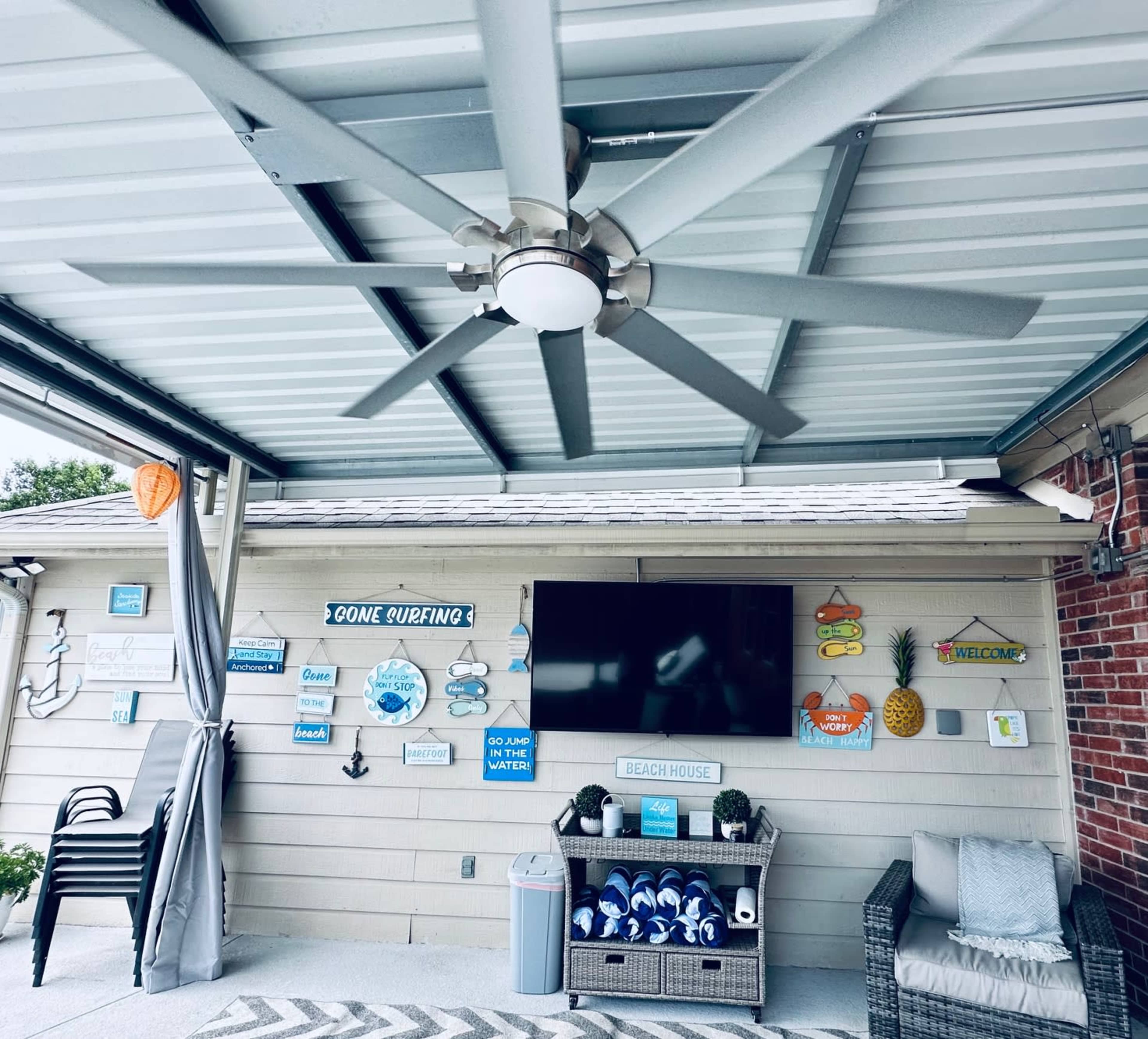 The image shows a covered patio area with a ceiling fan, a television mounted on the wall, and decor featuring various beach-themed signs and accessories.