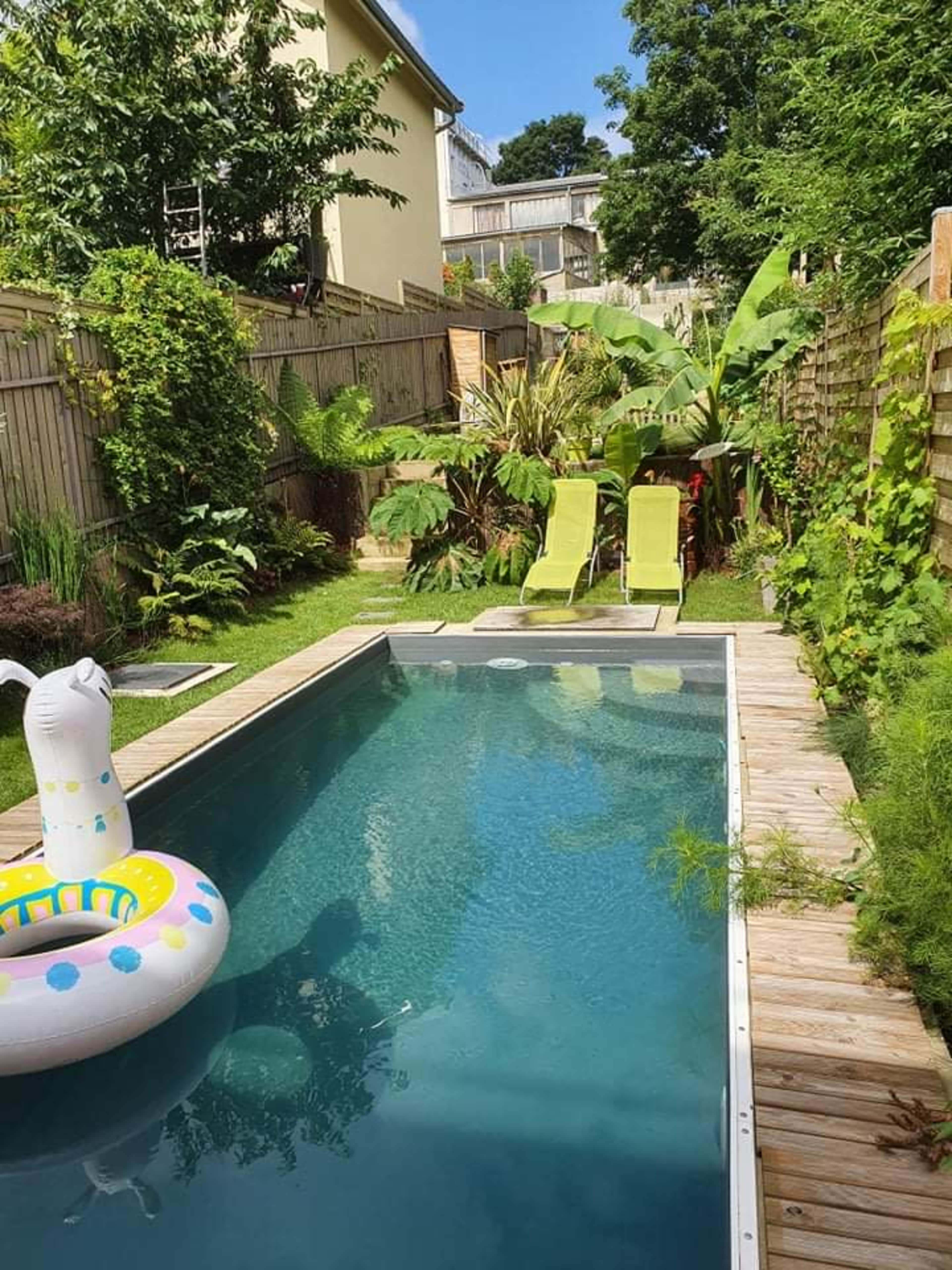 A backyard pool surrounded by greenery, featuring two lounge chairs and a colorful inflatable float in the water.