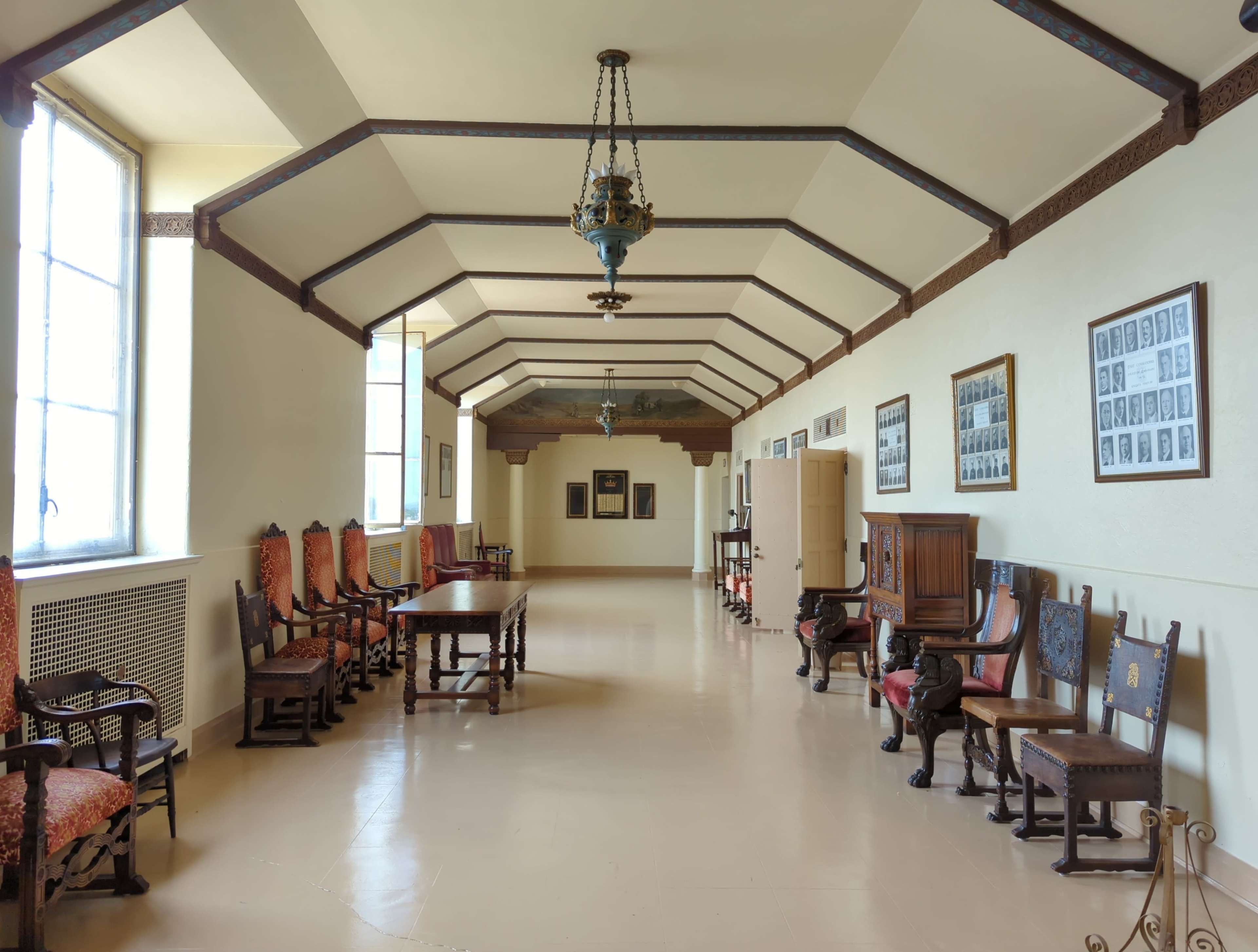 A long hallway with a row of chairs and tables, featuring framed photographs on the walls and large windows allowing natural light.
