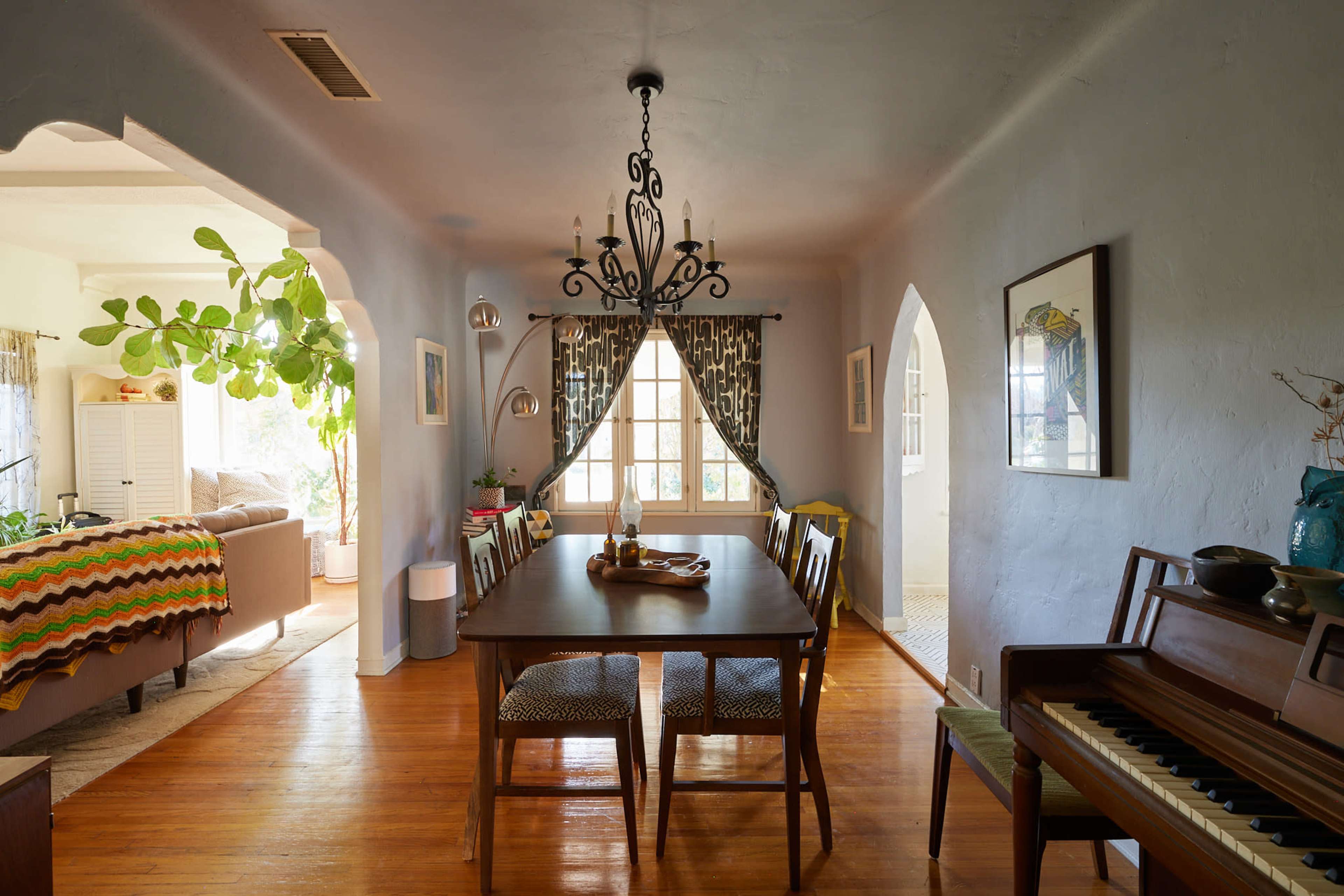 A dining room features a wooden table surrounded by chairs, with a chandelier overhead and a view into a living area with large windows.