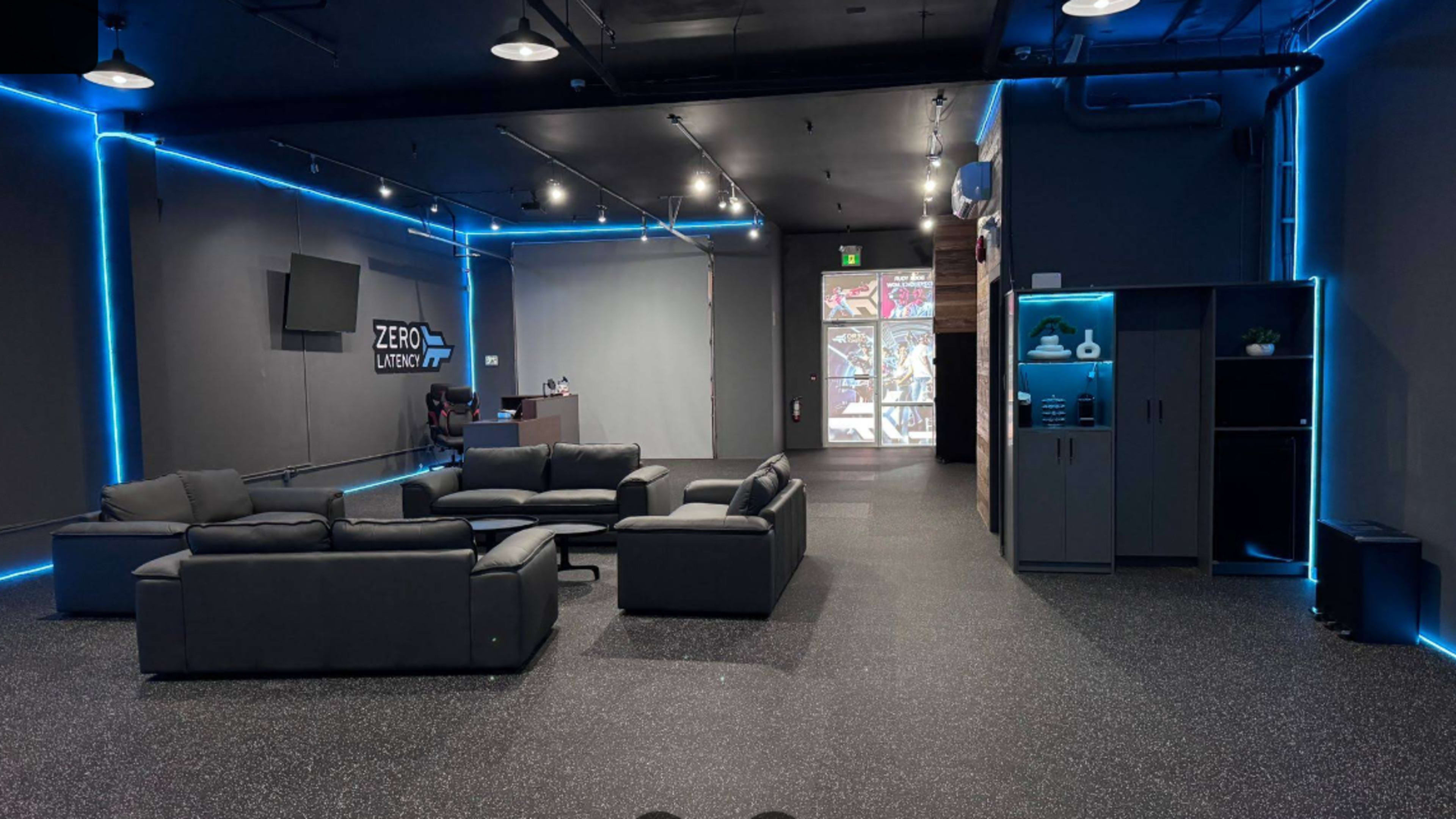 The image shows a modern, spacious lounge area with black furniture, neon blue lighting along the walls, and a reception desk in the background.