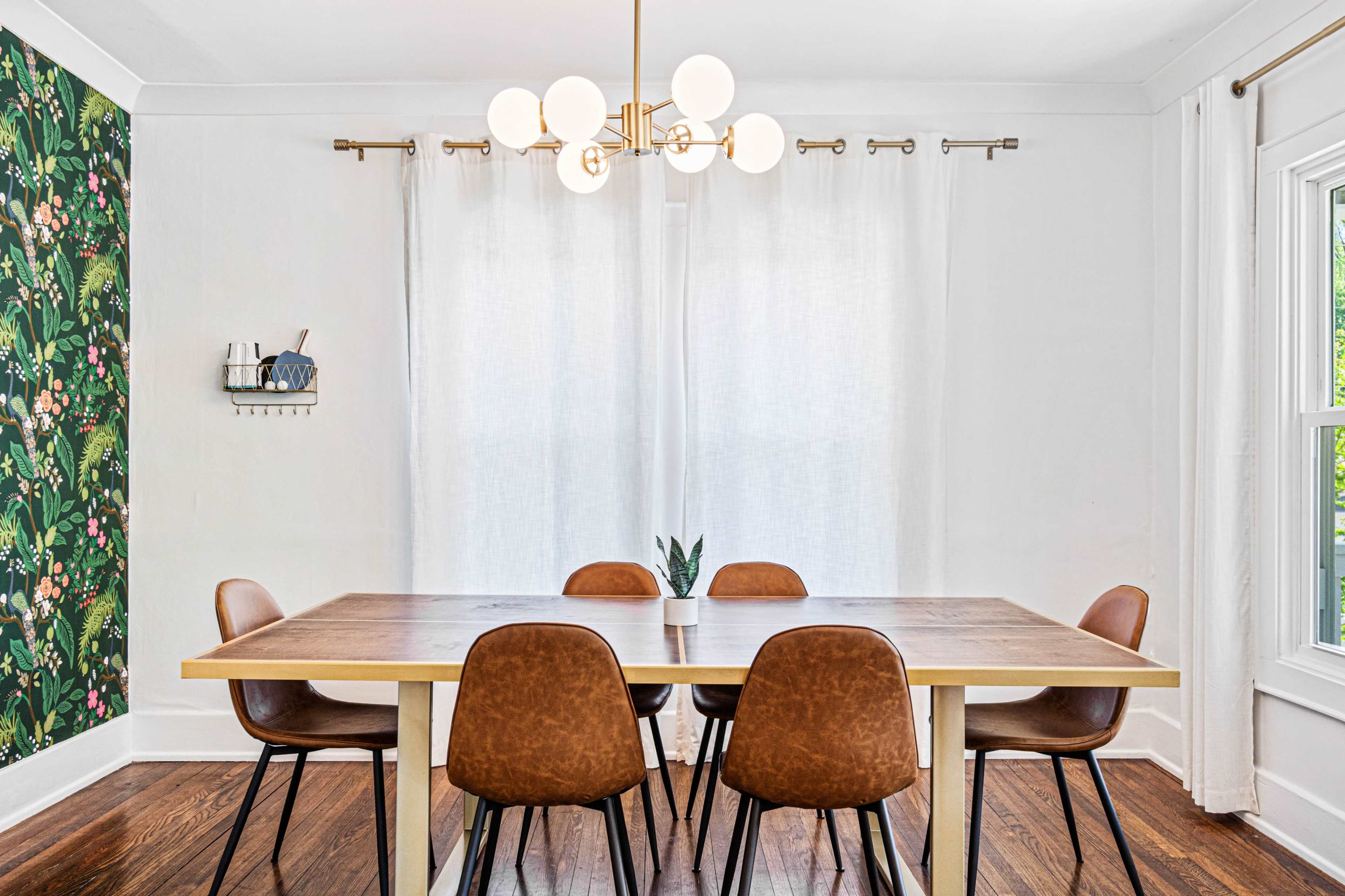 A dining room features a wooden table surrounded by six brown chairs, with a floral wallpaper accent on one wall and large windows covered by light curtains.