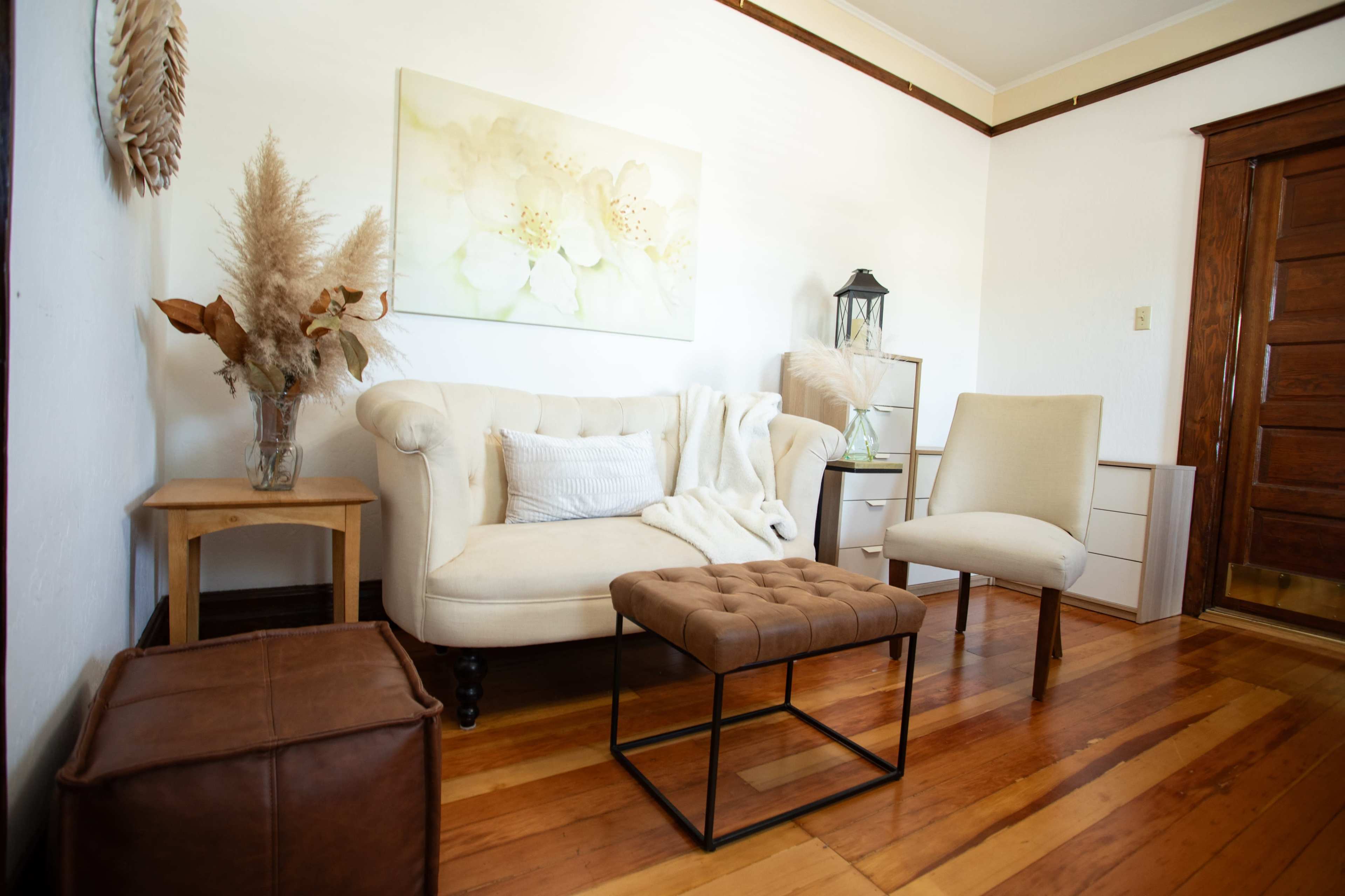 The image shows a cozy living room with a beige tufted sofa, a small wooden side table, a decorative armchair, and a brown leather ottoman, all situated on polished wooden floors.