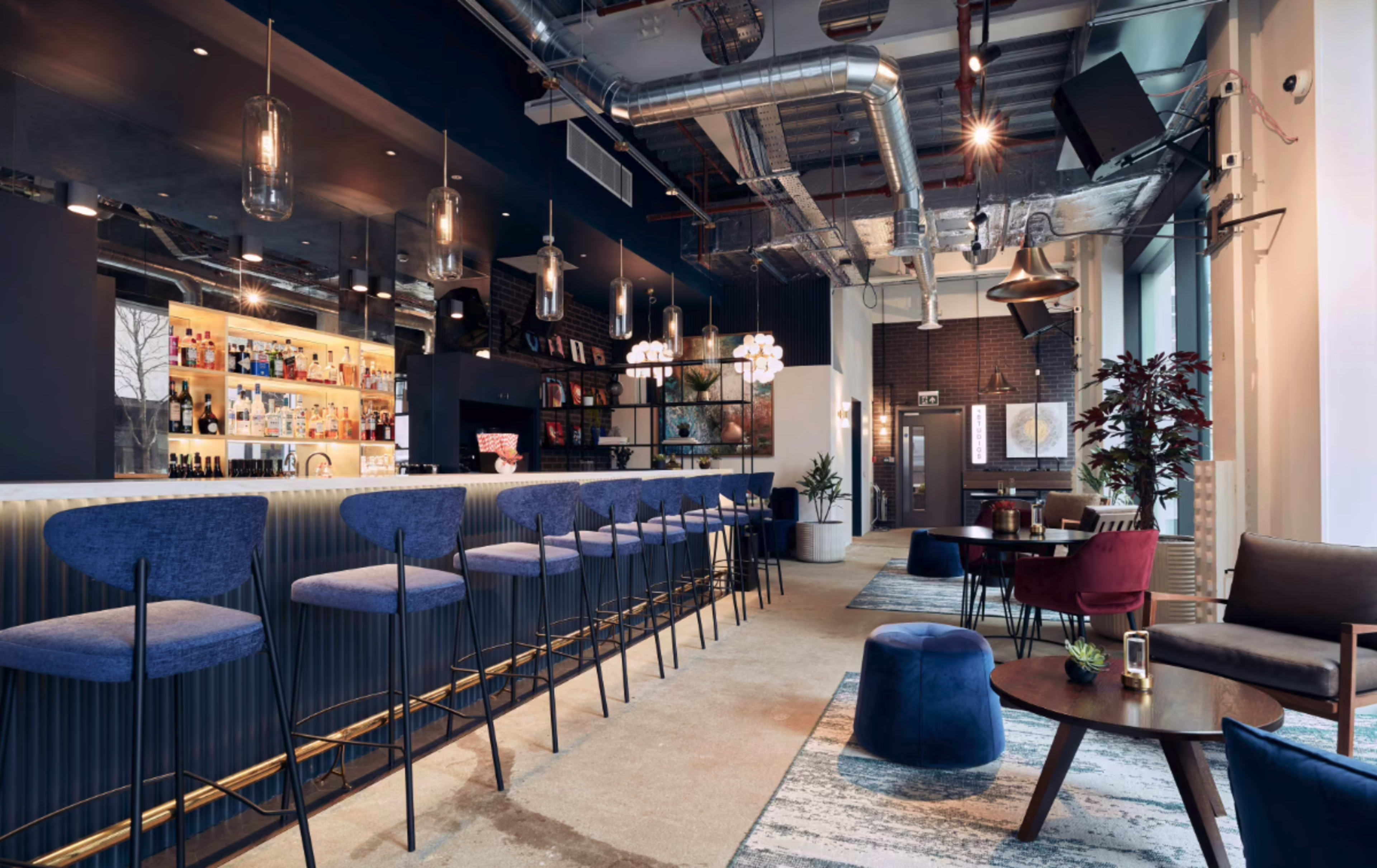 The image shows a modern bar area with a long counter, several stylish high stools, and a mix of seating arrangements including lounge chairs and tables.