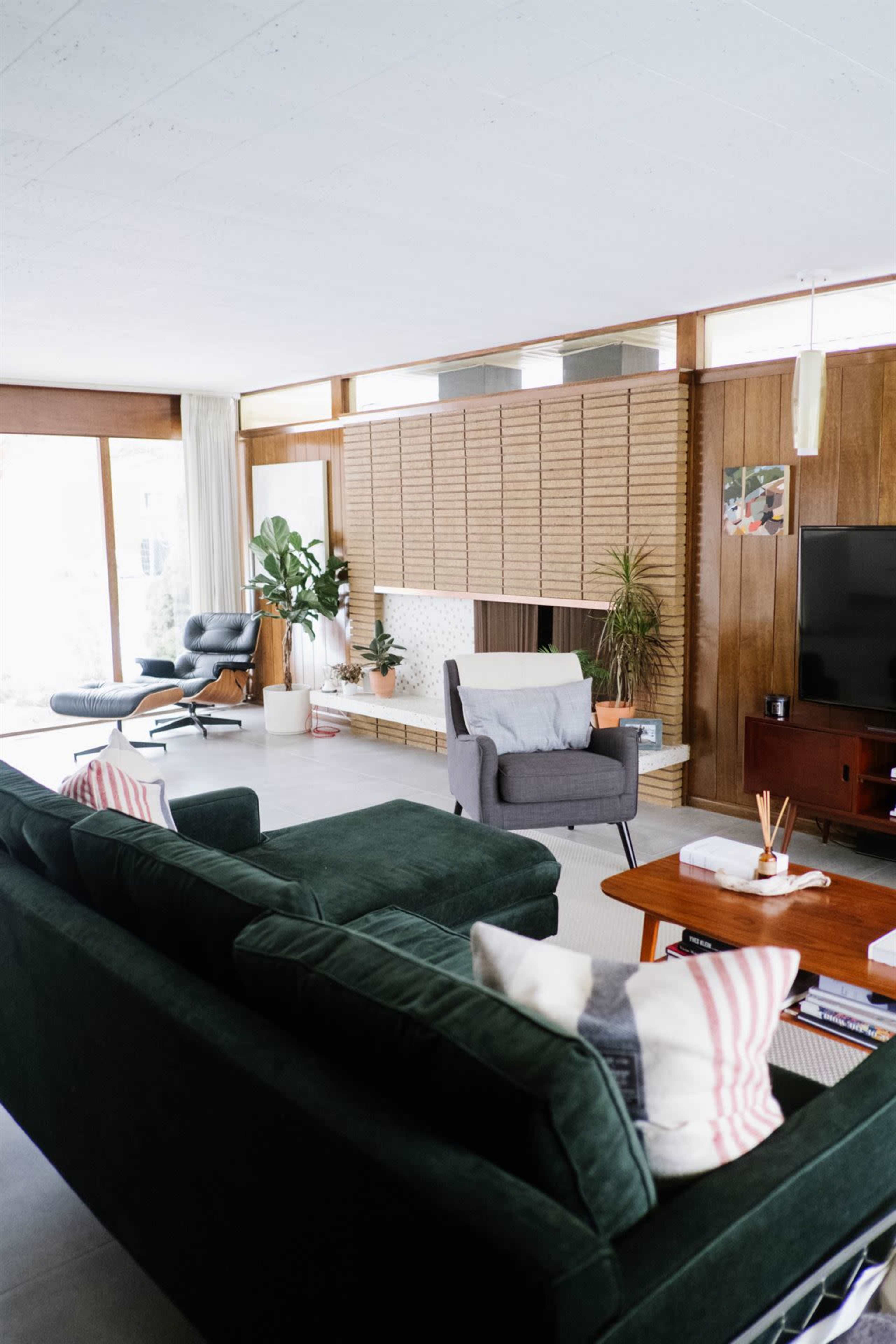 A modern living room features a green sofa, a gray armchair, and a large wall of wooden paneling with a fireplace, complemented by plants and a television.