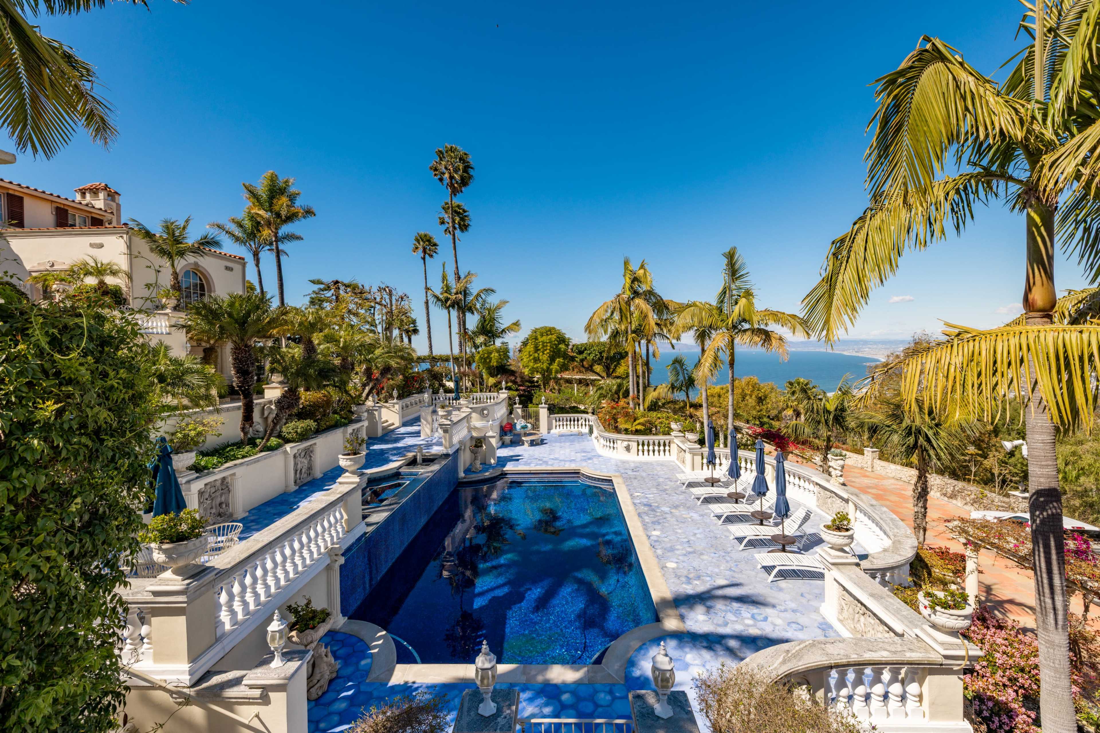 The image features a luxurious villa with a blue swimming pool surrounded by palm trees and landscaped gardens, overlooking a body of water.