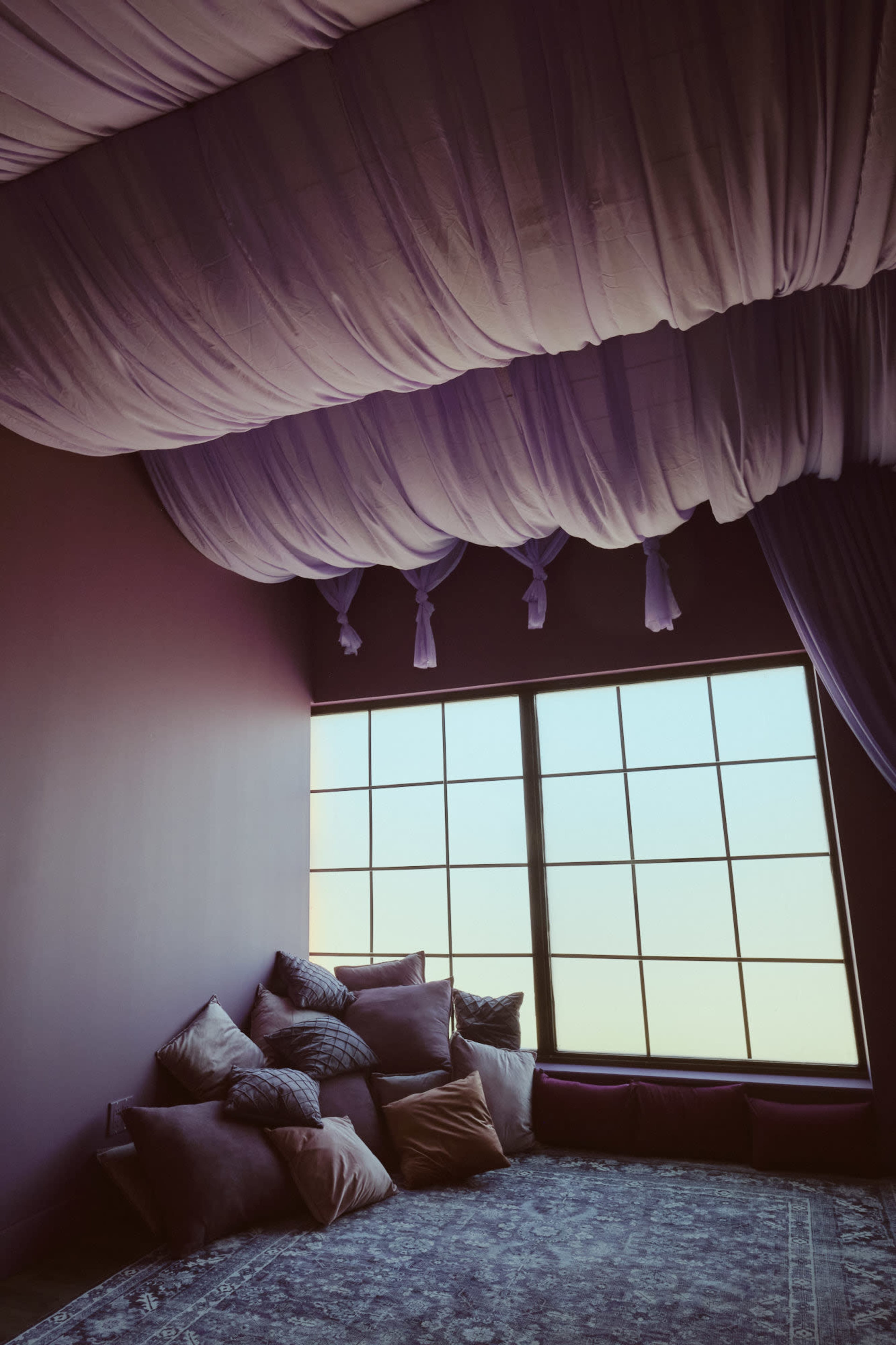 The image shows a cozy corner of a room with a large window, plush pillows arranged on the floor, and draped fabric on the ceiling.