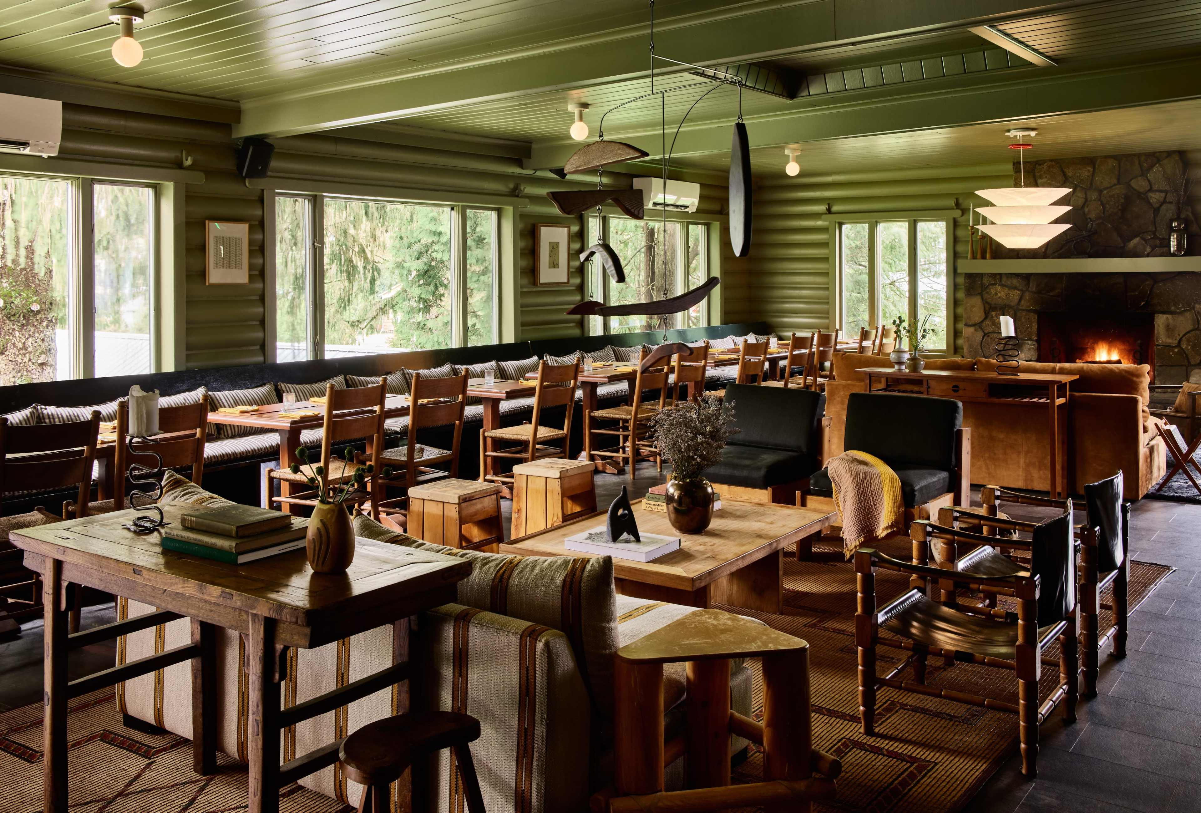 A spacious dining area with wooden tables and chairs, large windows allowing natural light, and a stone fireplace at one end.