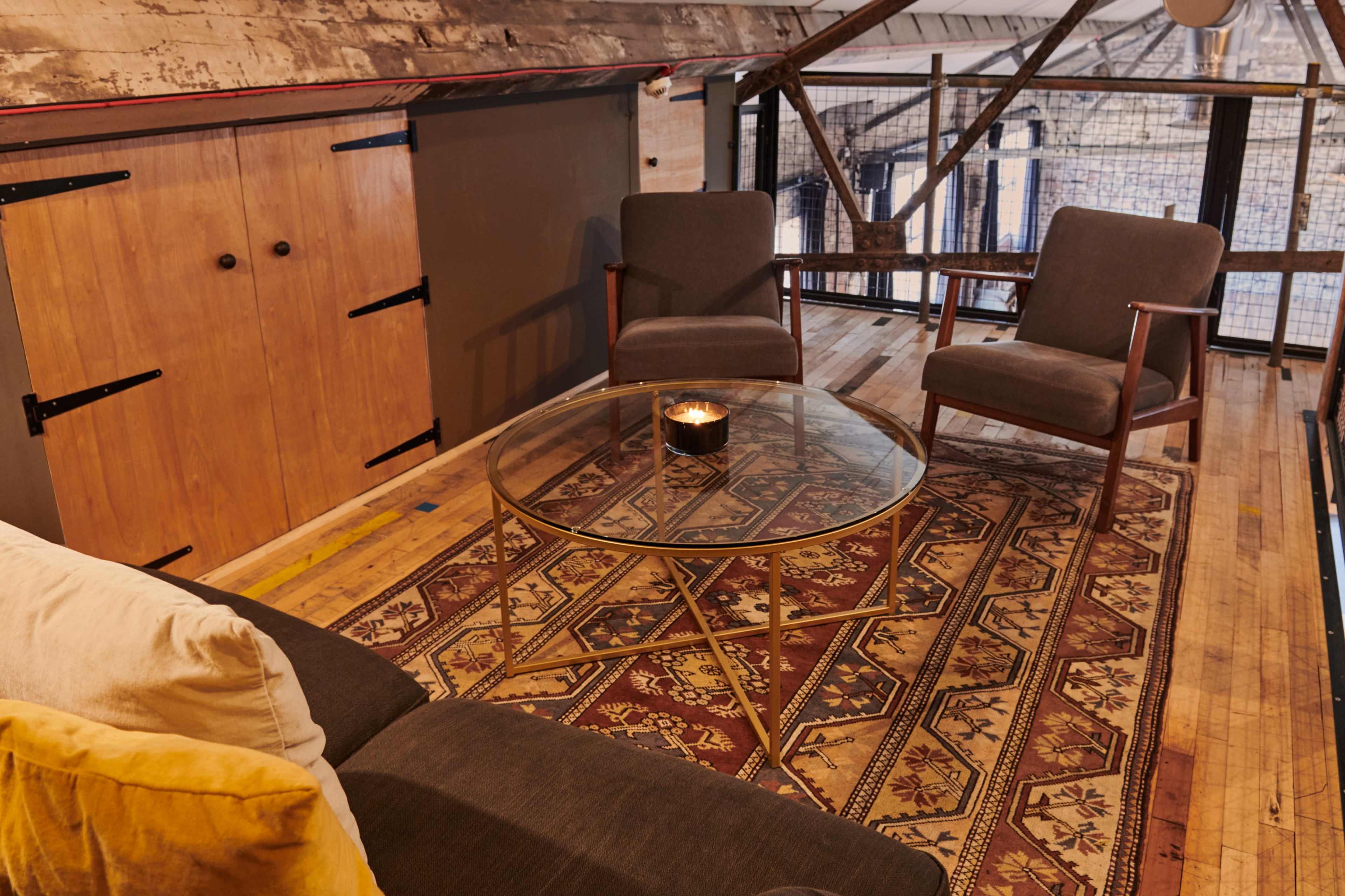 A cozy loft living space featuring two armchairs, a glass coffee table on a patterned rug, and wooden walls with storage cabinets.